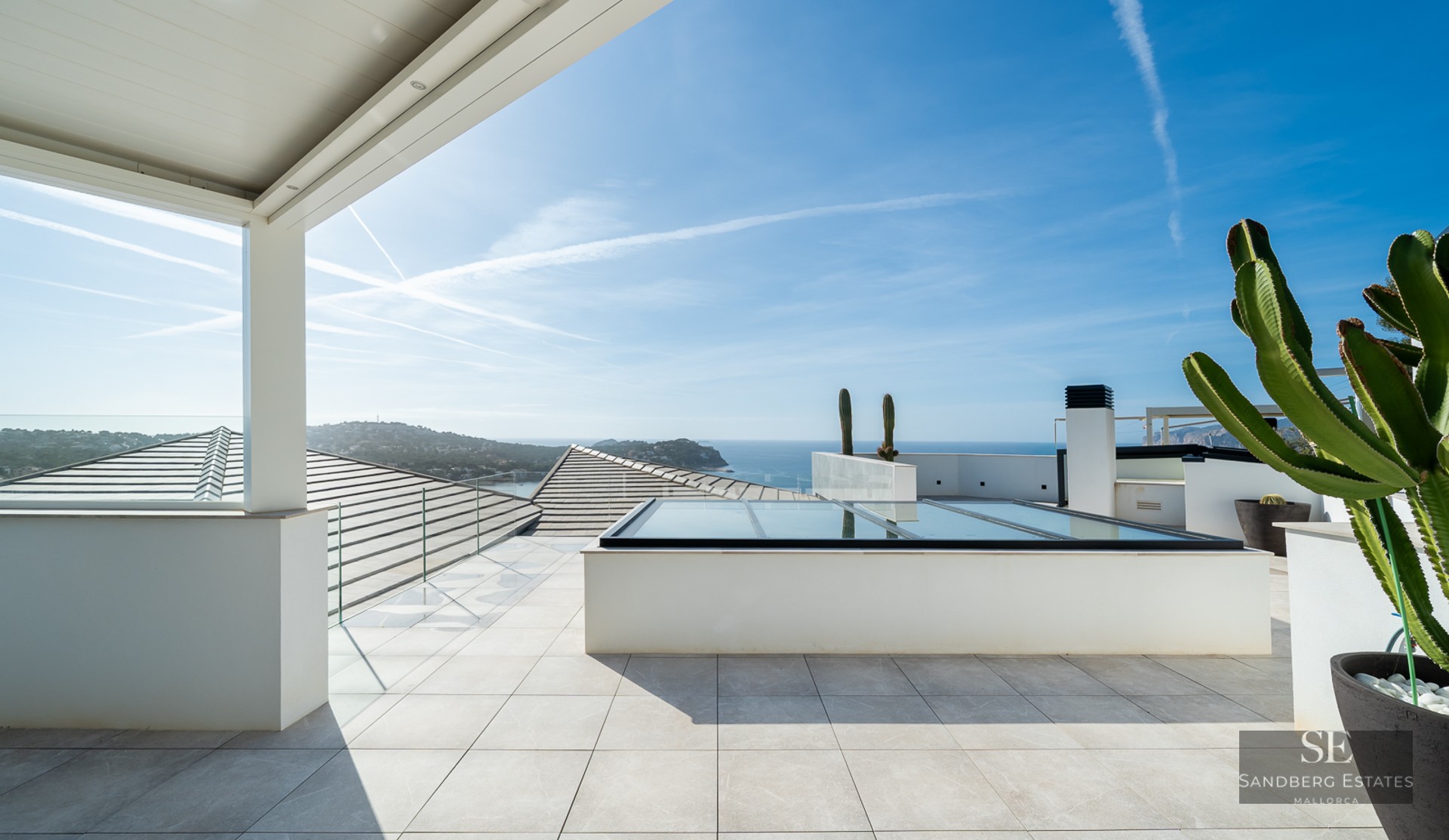 Bright white rooftop terrace with glass railings overlooking the sea and sky with large cacti in pots.