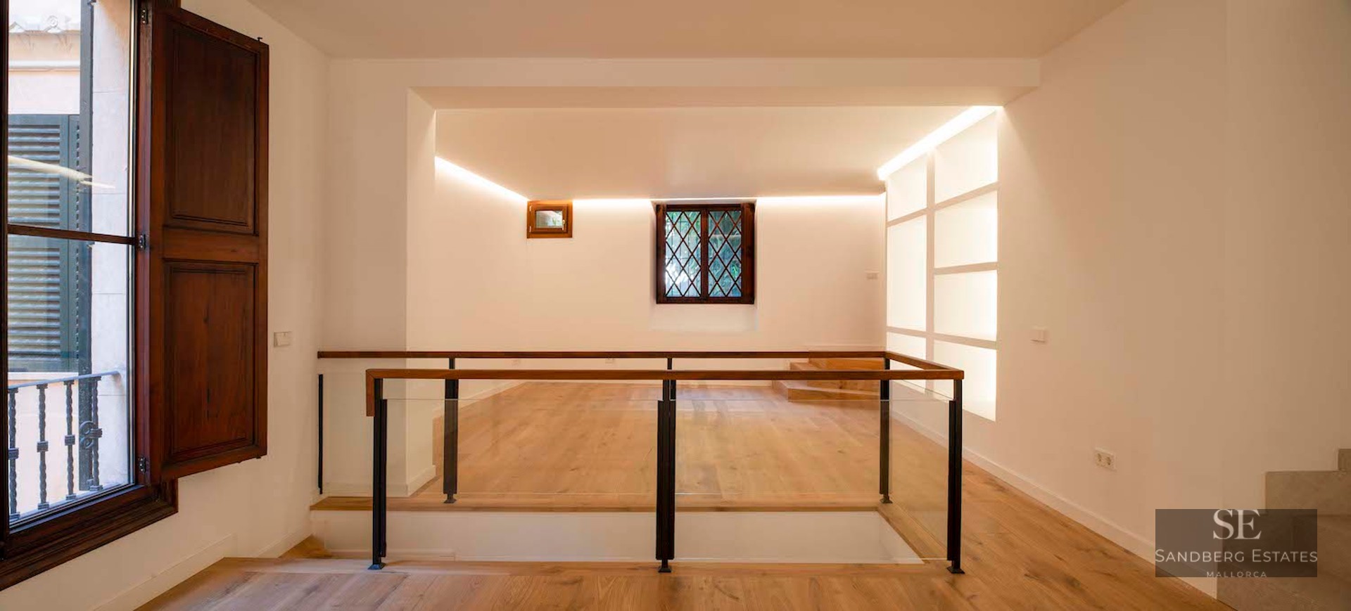 A modern indoor landing with light oak flooring, glass railings, and warm cove lighting against white walls.