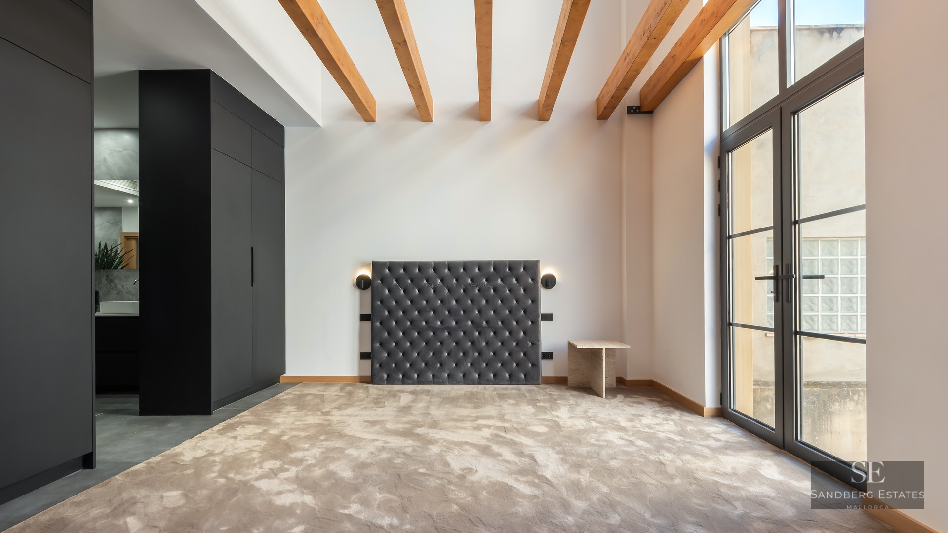 Bedroom featuring a grey velvet tufted headboard, wooden ceiling beams, black wardrobe, and large glass doors.
