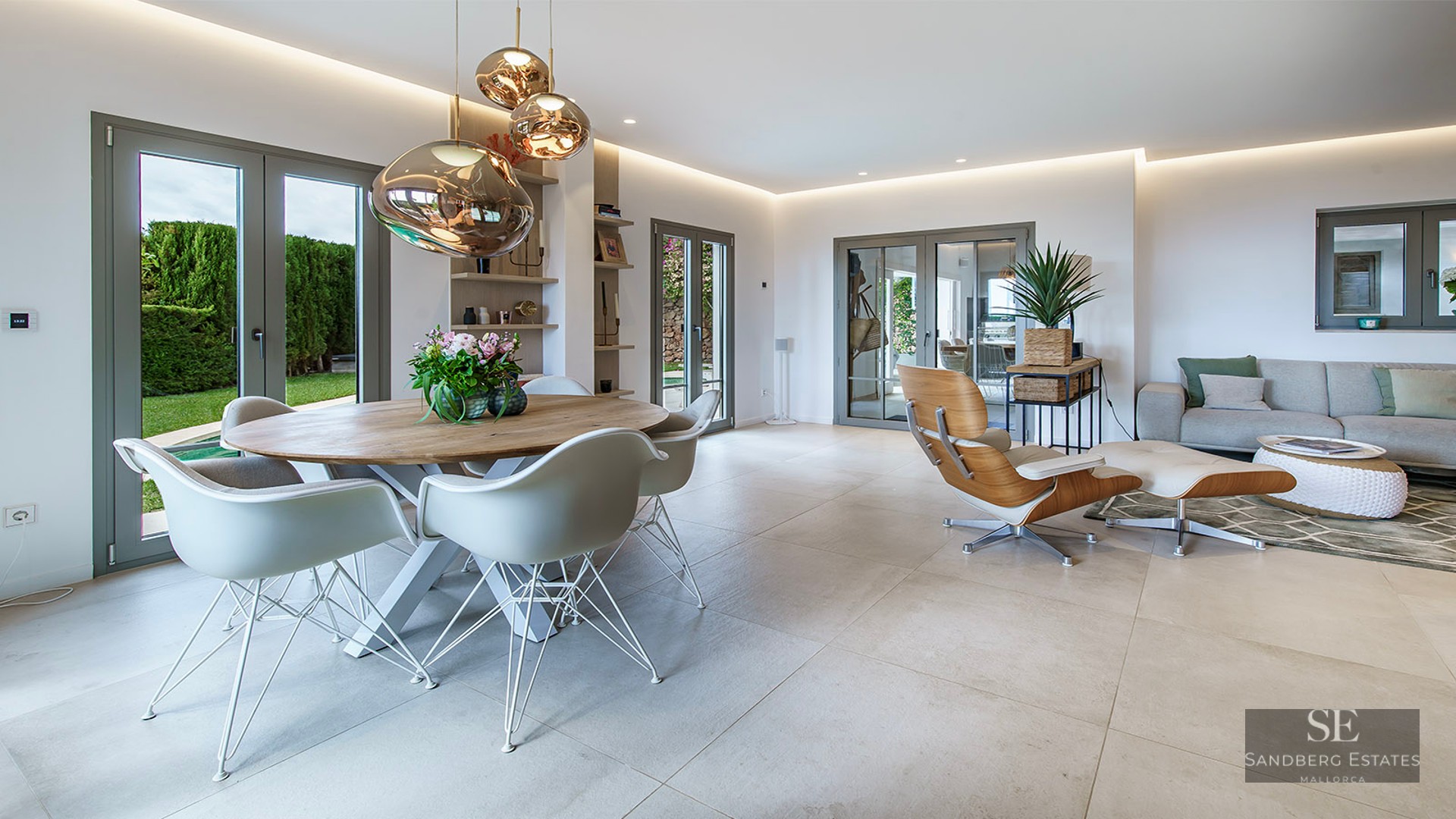 Modern open-plan living and dining room with designer furniture, tile floors, and glass doors to a green garden.