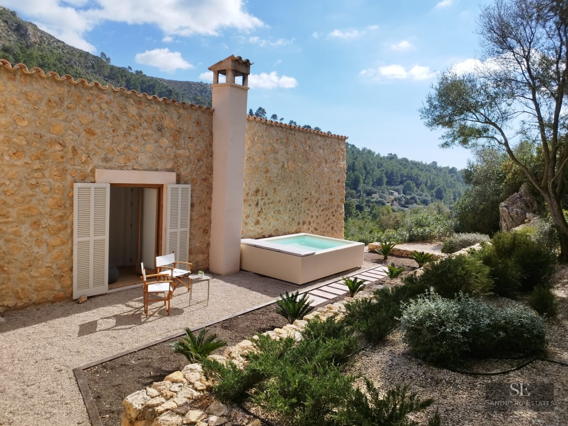 Stone house featuring an outdoor jacuzzi, wooden chairs on gravel, and mountain views under a clear sky.