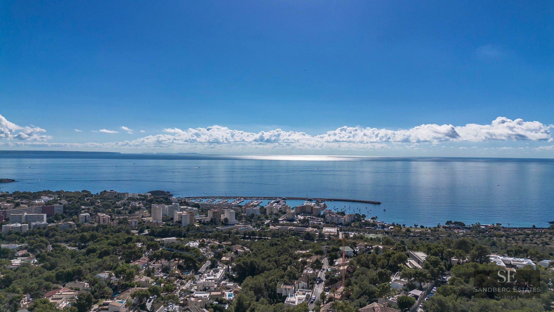 Vue aérienne d'une ville côtière avec marina, arbres verts et mer Méditerranée sous un ciel bleu clair.