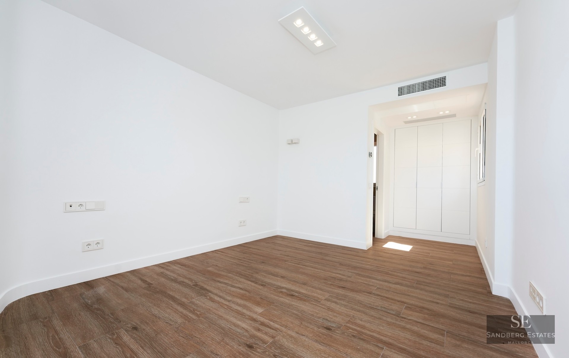 Empty modern room with white walls, wood-look flooring, and white built-in wardrobes in the background.
