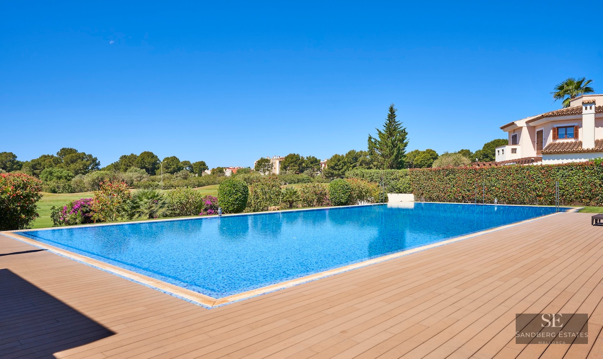 Large blue swimming pool with a wooden deck, surrounded by green hedges and a Mediterranean villa under a clear sky.