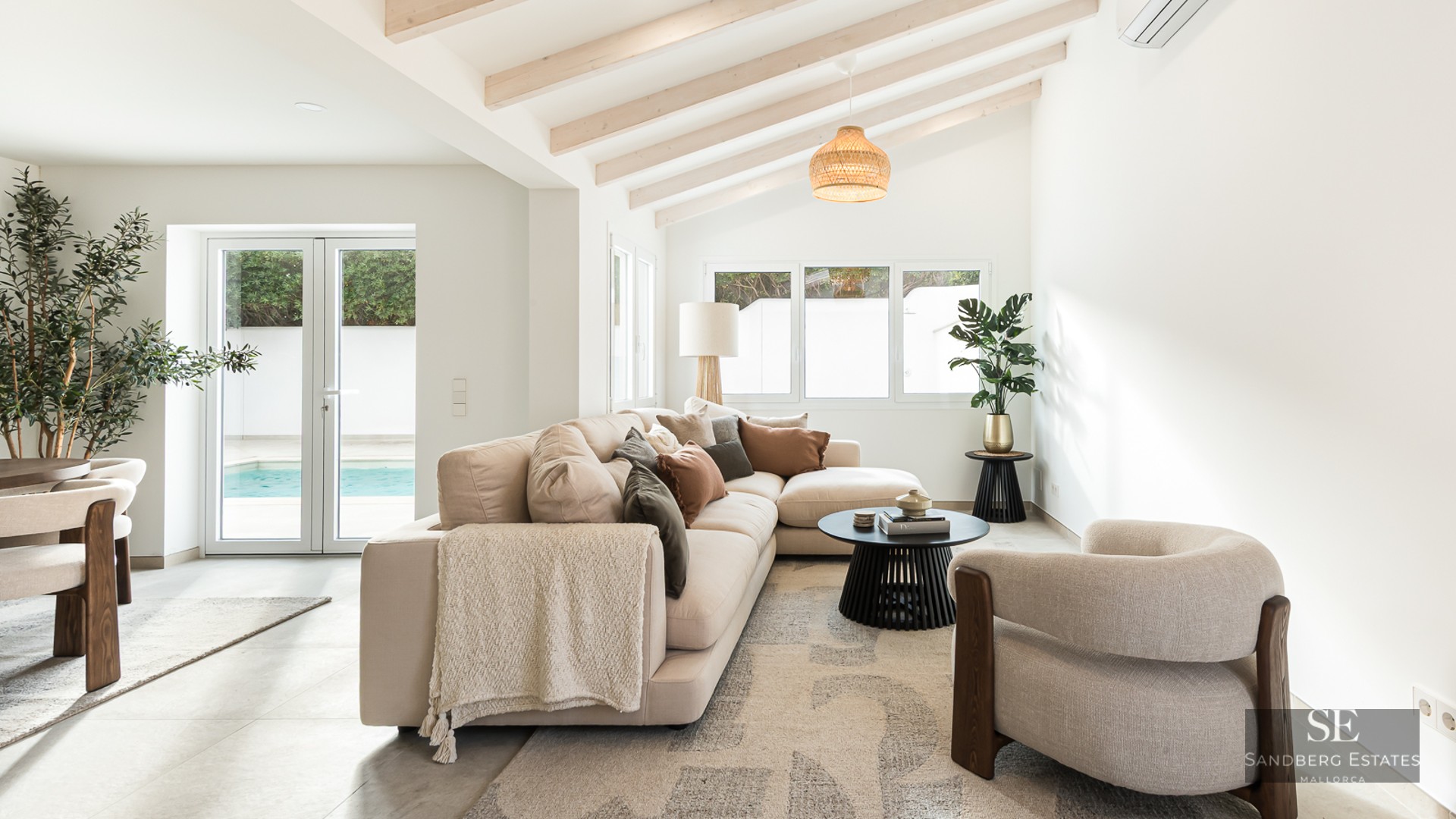 Bright living room with beige sectional sofa, wooden ceiling beams, and glass doors opening to a swimming pool.