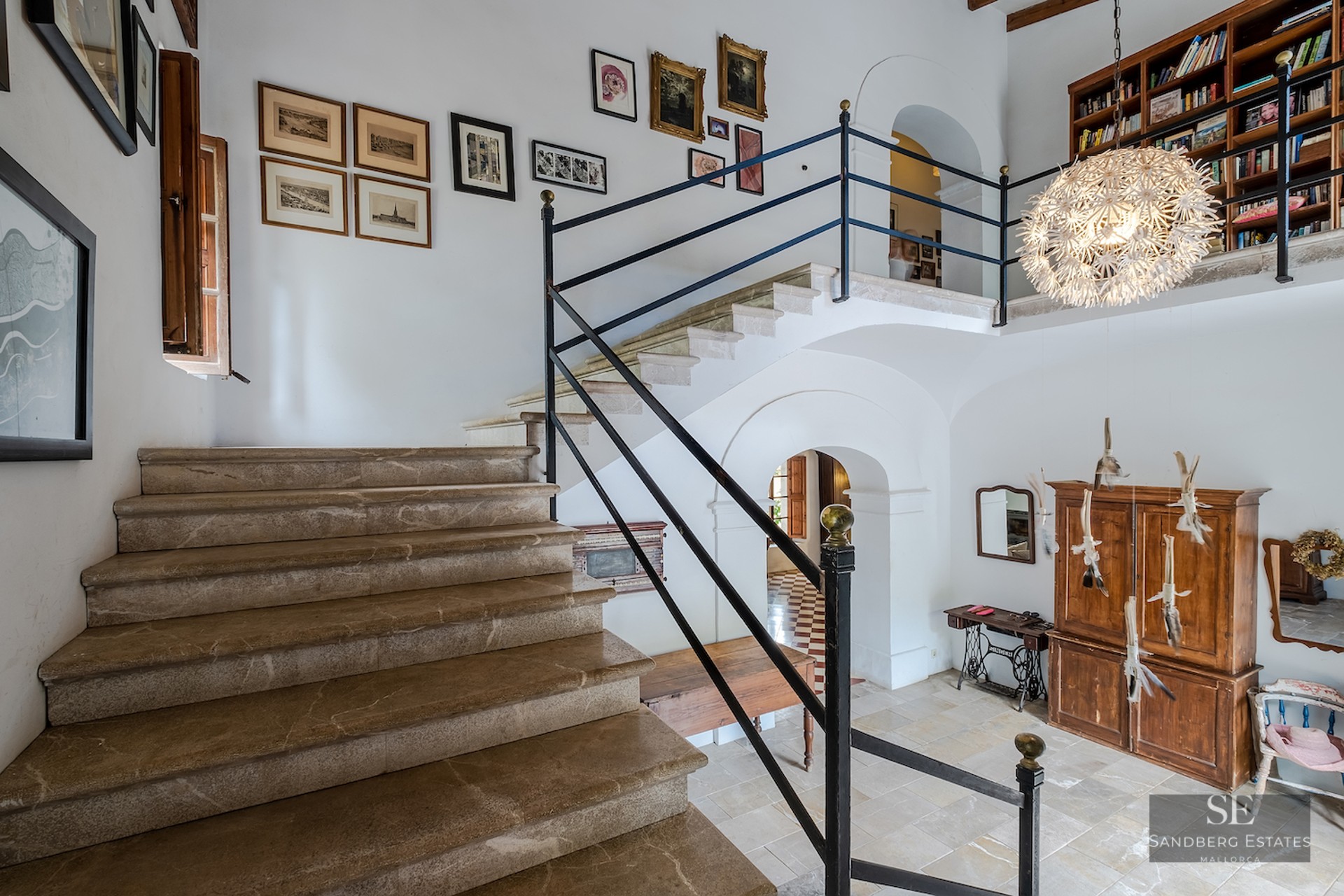 Grand entryway featuring a stone staircase, white walls adorned with art, and a mezzanine library level.