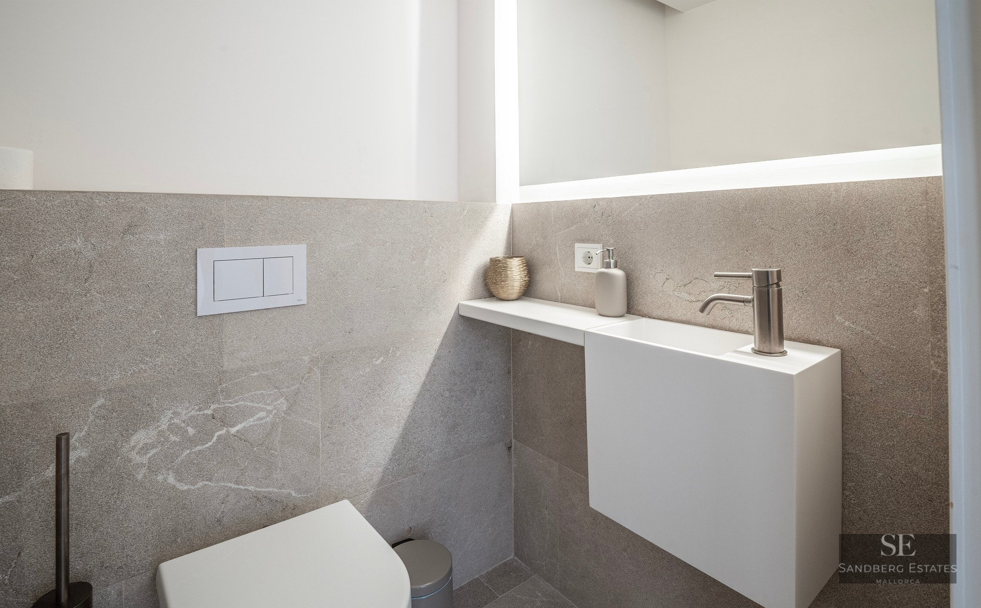 Modern powder room with textured stone tiles, a white floating sink, wall-mounted toilet, and a large backlit mirror.