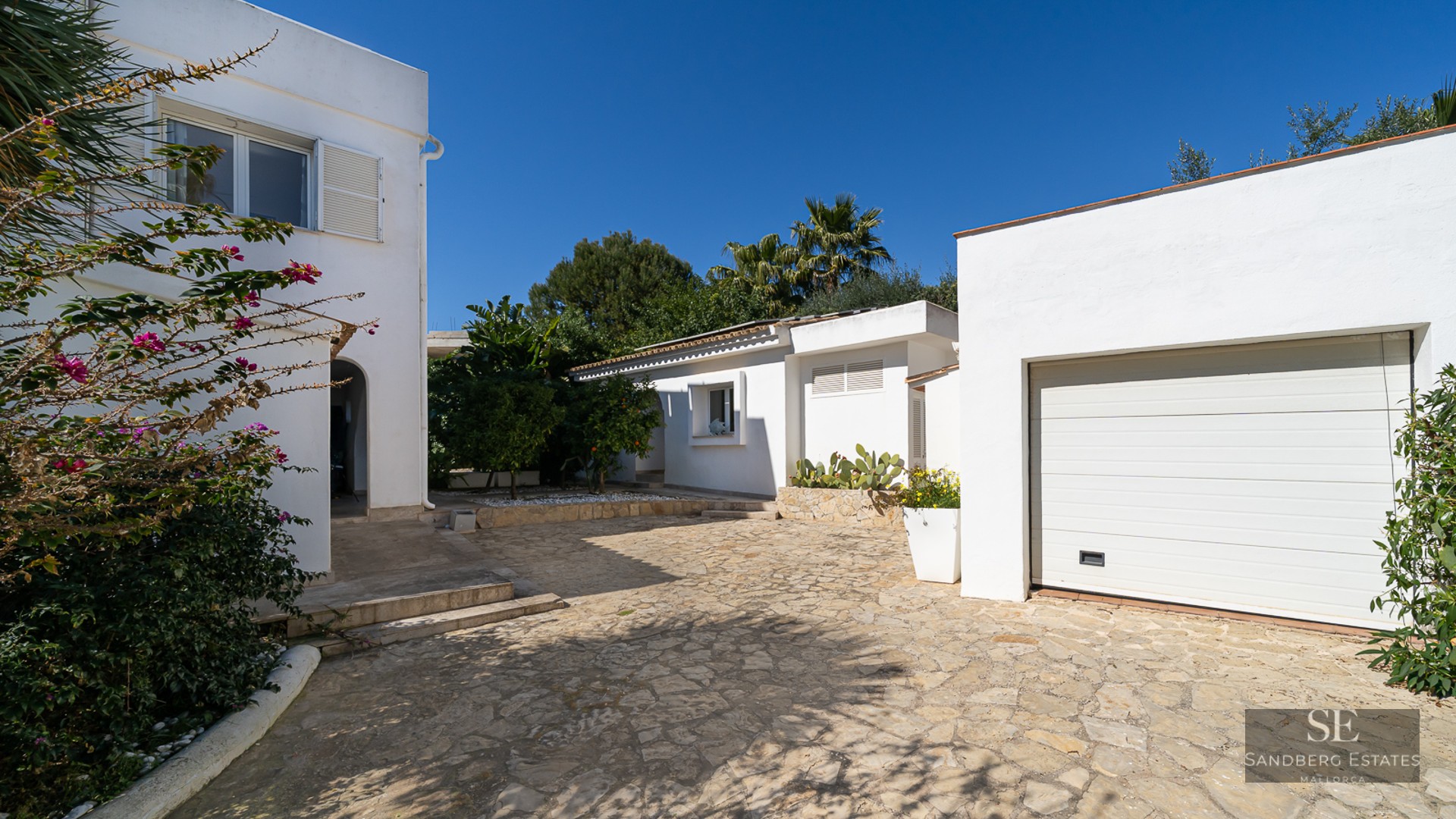 White Mediterranean villa with a stone-paved driveway, garage, and lush greenery under a clear blue sky.