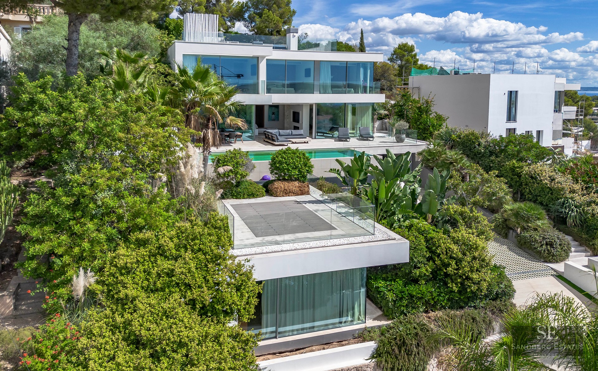 Modern multi-level white luxury villa with expansive glass walls, a turquoise pool, and dense Mediterranean landscaping.