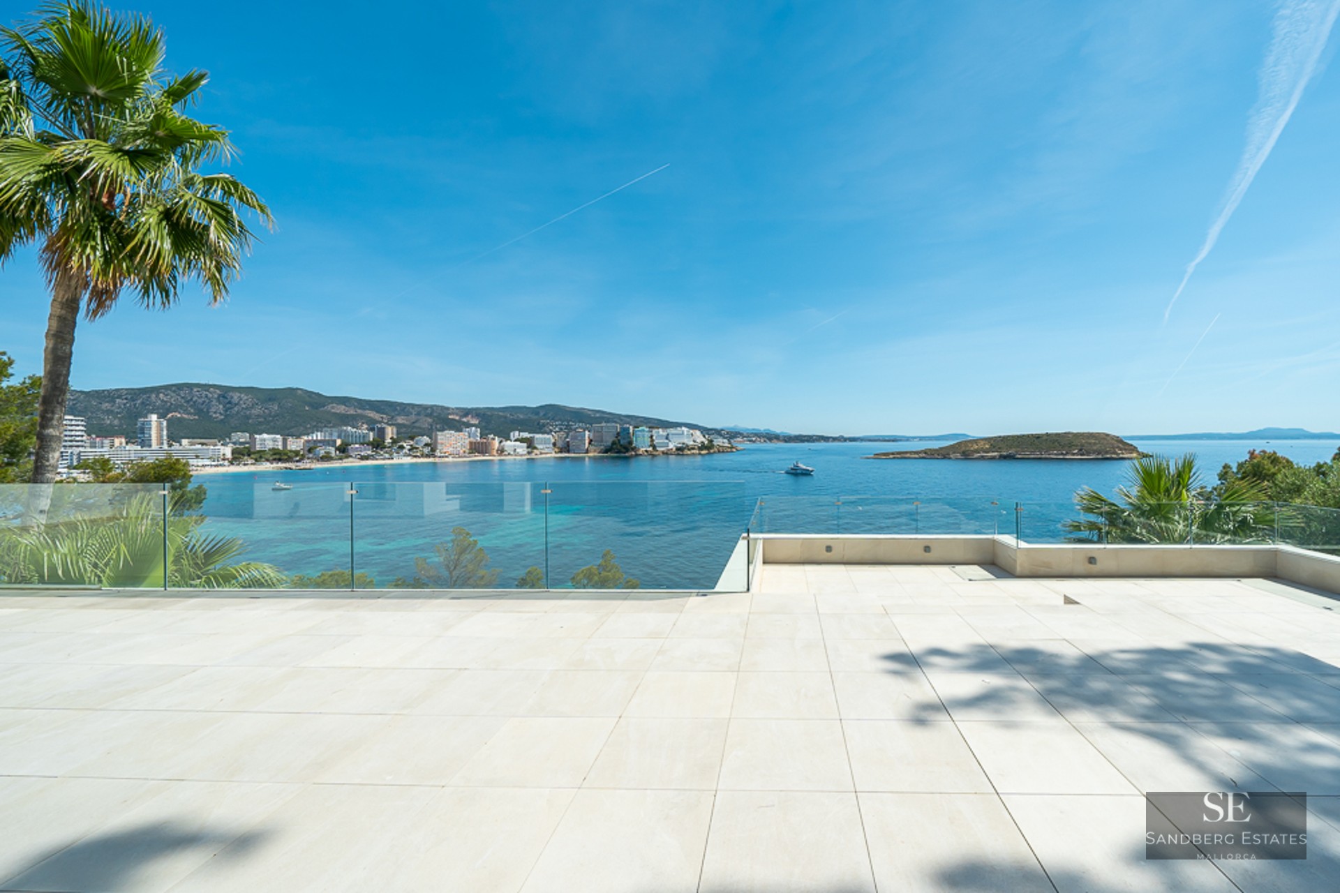 Large white tiled terrace with glass railings overlooking a blue bay, mountains, and coastal town.