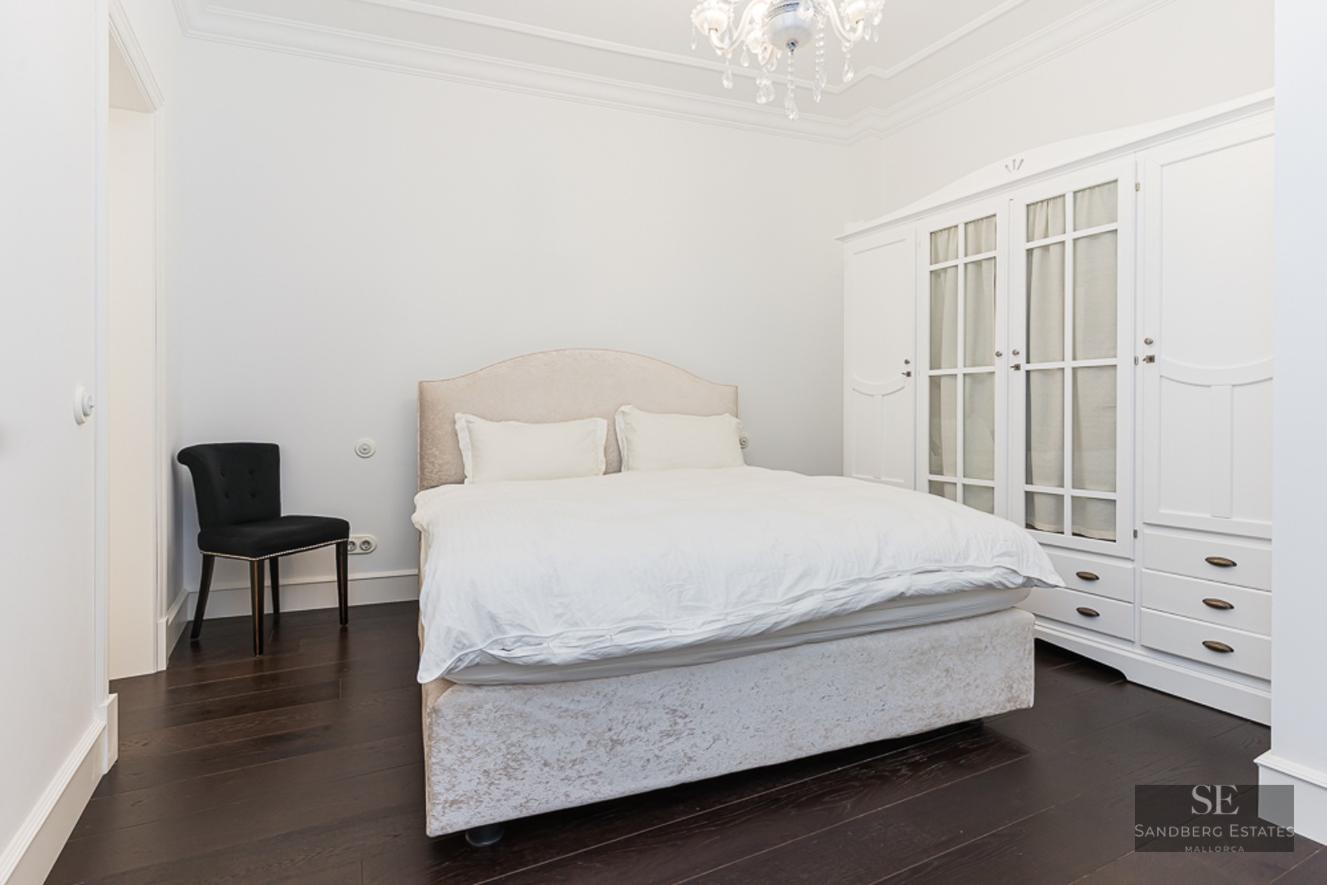 Bright bedroom featuring a beige bed, large white wardrobe, black chair, and crystal chandelier on dark wood floors.