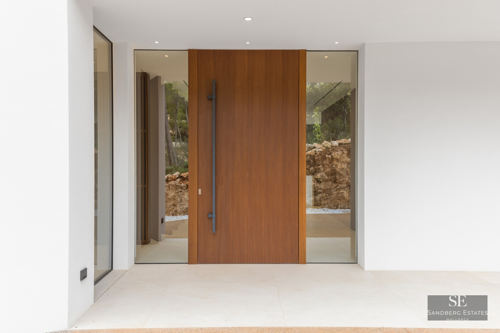 Large wooden pivot door with glass side panels flanked by smooth white walls and minimalist lighting.