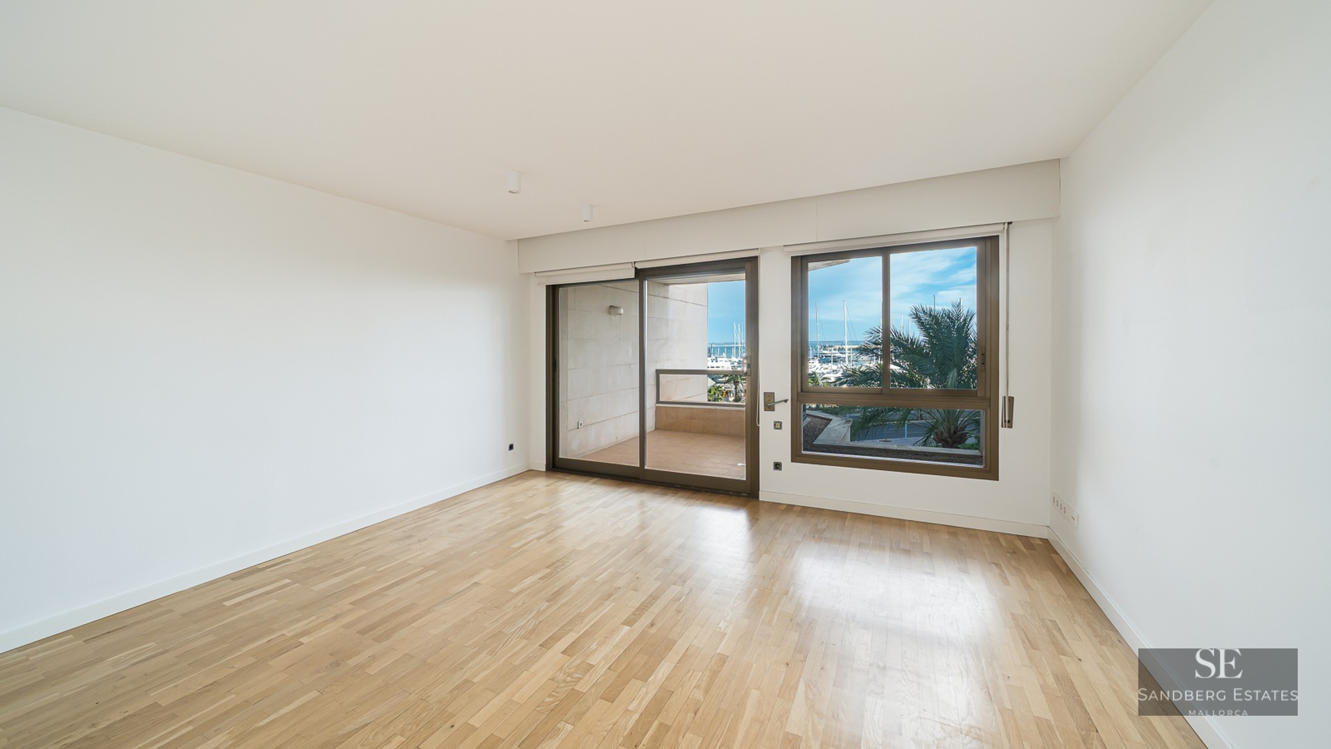 Empty modern room with light wood floors and large windows overlooking a balcony and a marina with palm trees.
