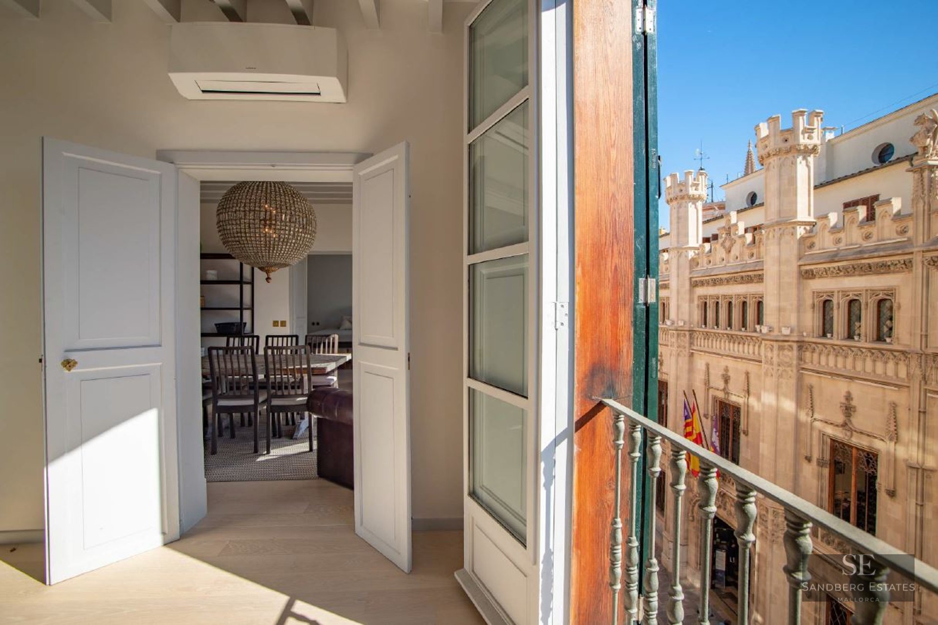 View from an open balcony door looking out at a historic Gothic stone building under a clear blue sky.