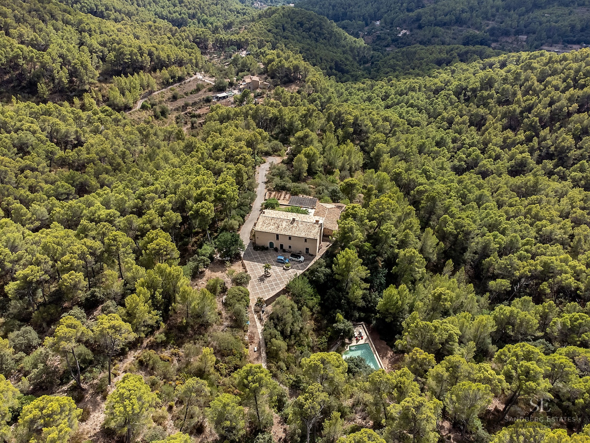 Aerial view of a large stone villa with a pool, surrounded by a dense green pine forest on a hillside.