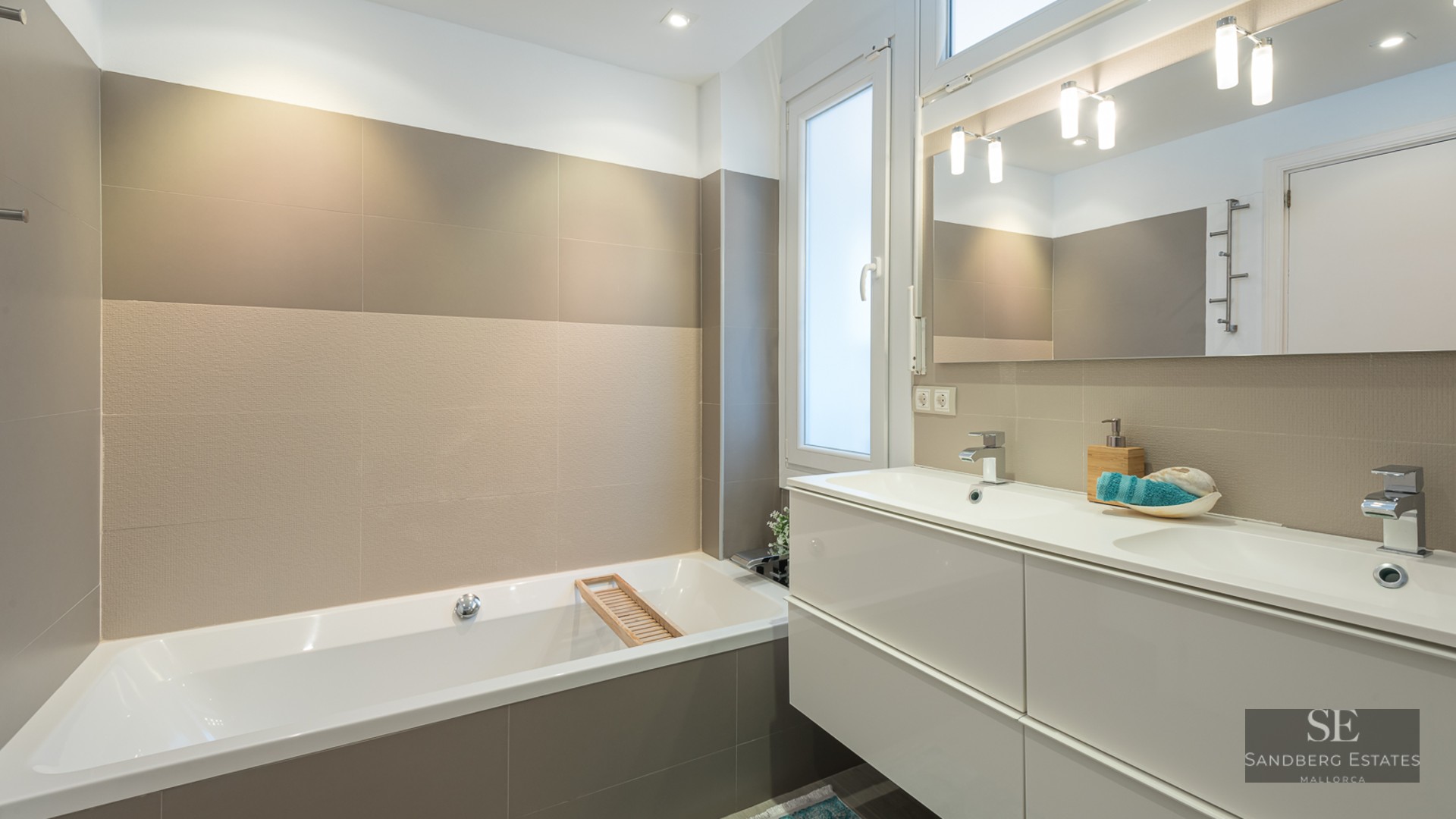 A contemporary bathroom with a white bathtub, a double-sink vanity, large beige tiles, and a wide mirror.