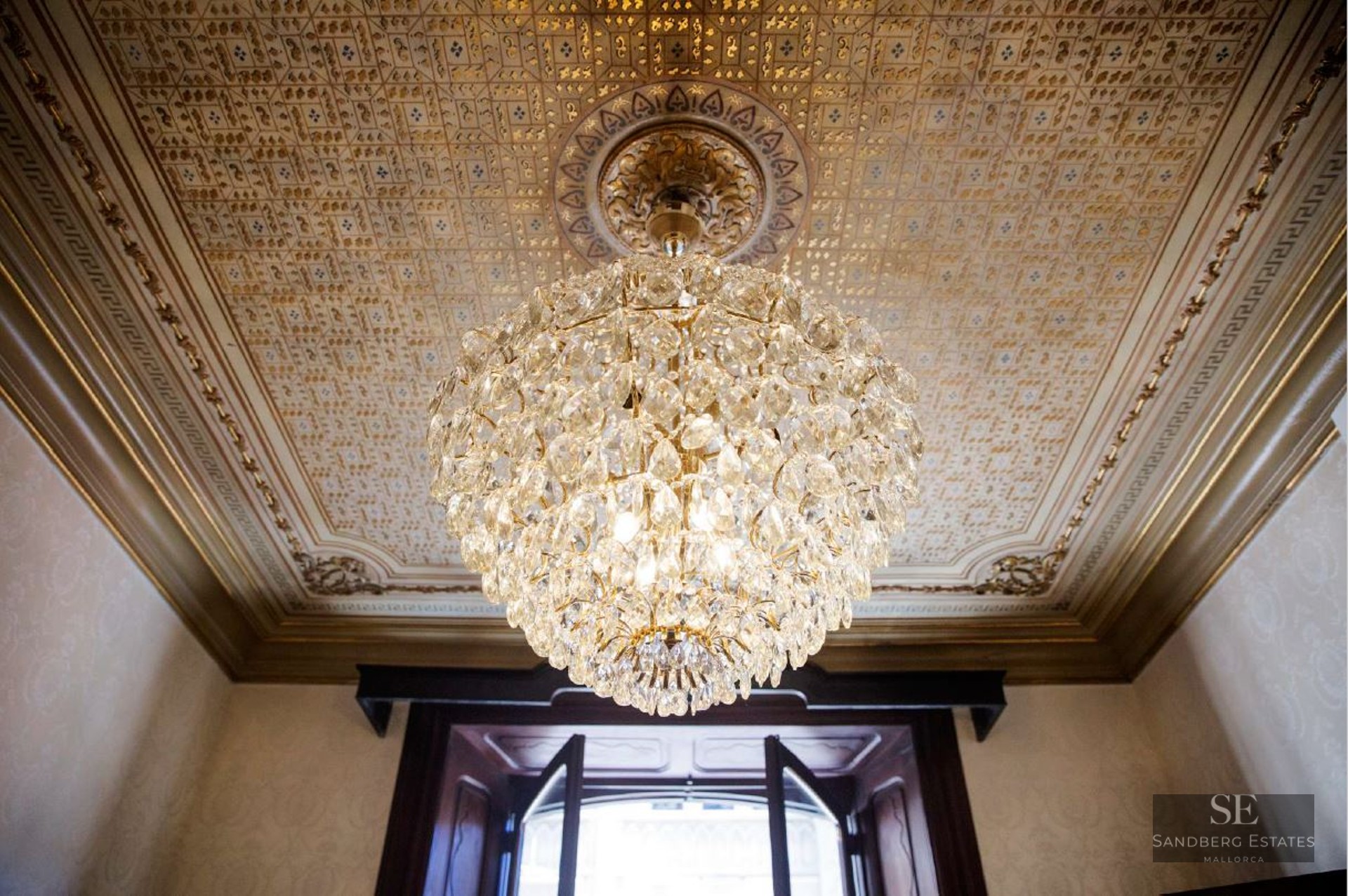 A large crystal chandelier hanging from a highly decorative gold-patterned ceiling with ornate moldings.