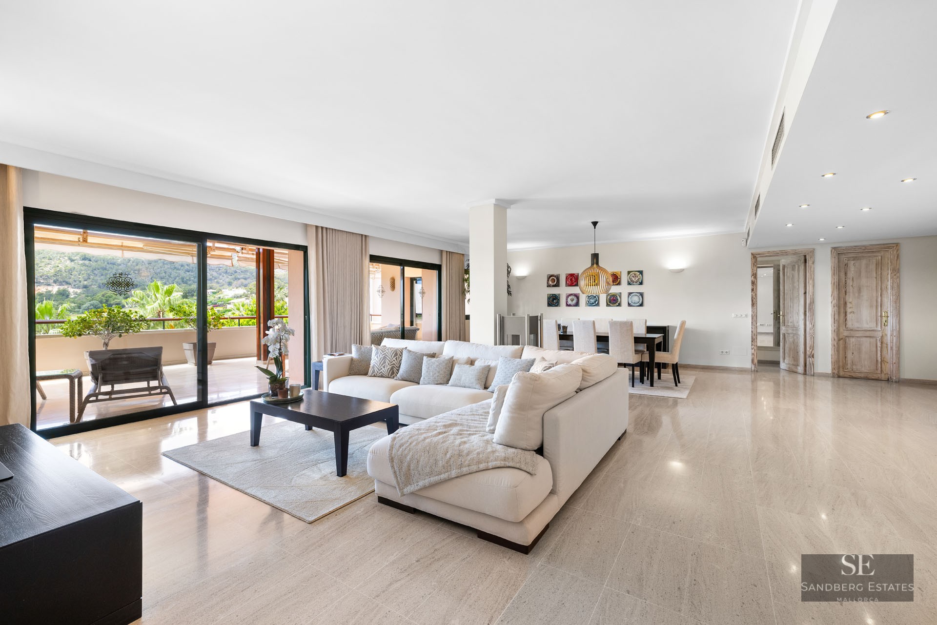 Salon spacieux avec canapé blanc en L, sol en marbre et grandes baies vitrées ouvrant sur une terrasse paysagée.