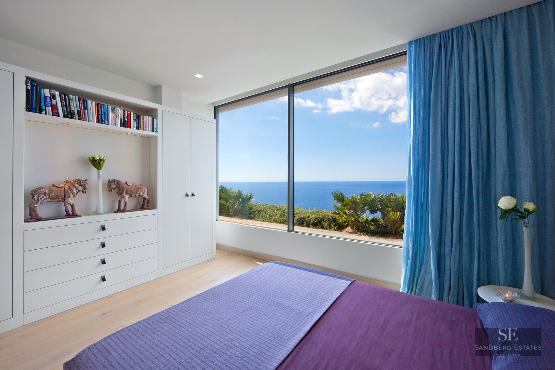 Modern white bedroom featuring a purple bed, large floor-to-ceiling window overlooking the blue sea and sky.