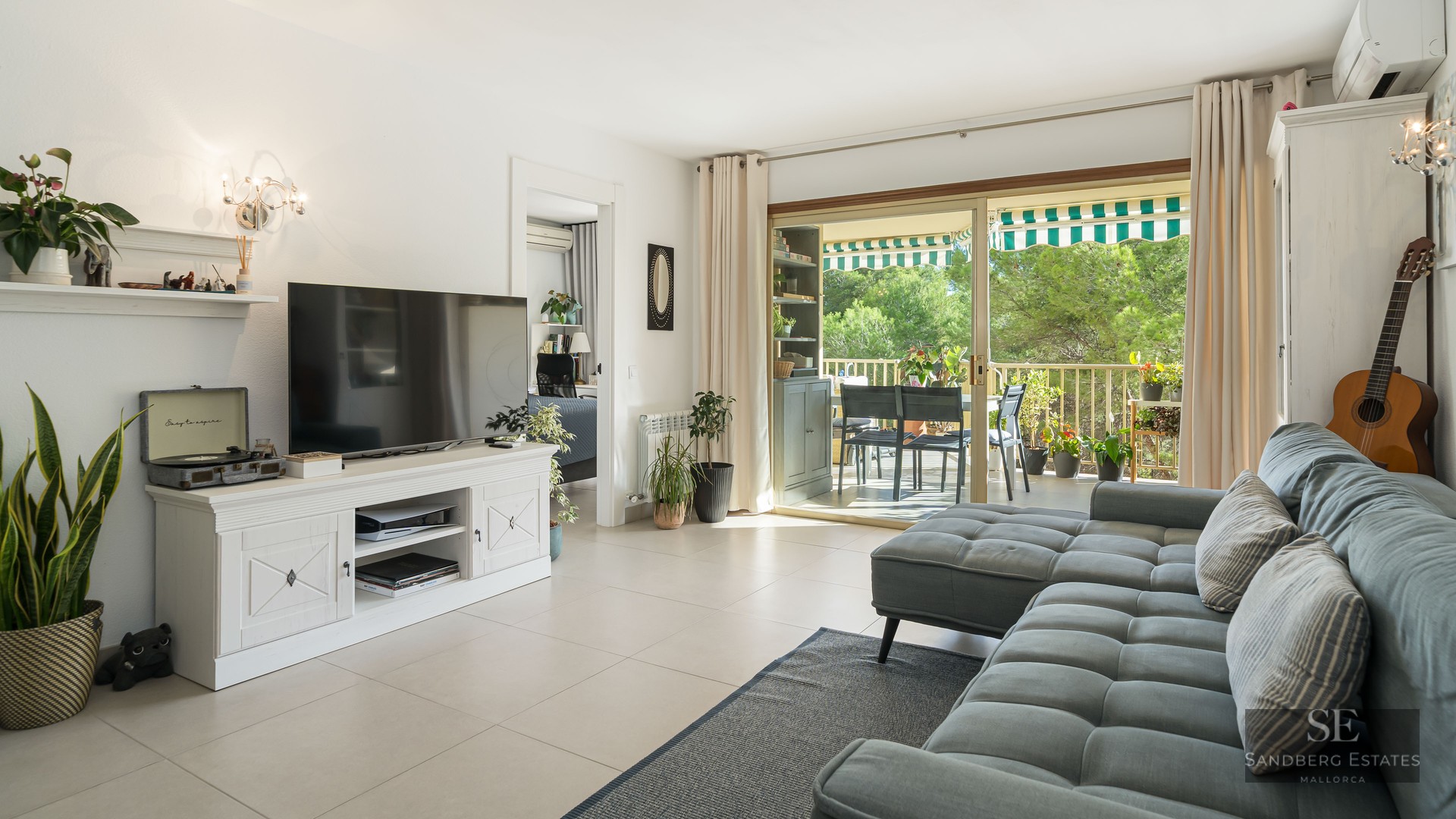 Salon spacieux avec canapé gris, télévision sur meuble blanc et baies vitrées ouvrant sur une terrasse verdoyante.
