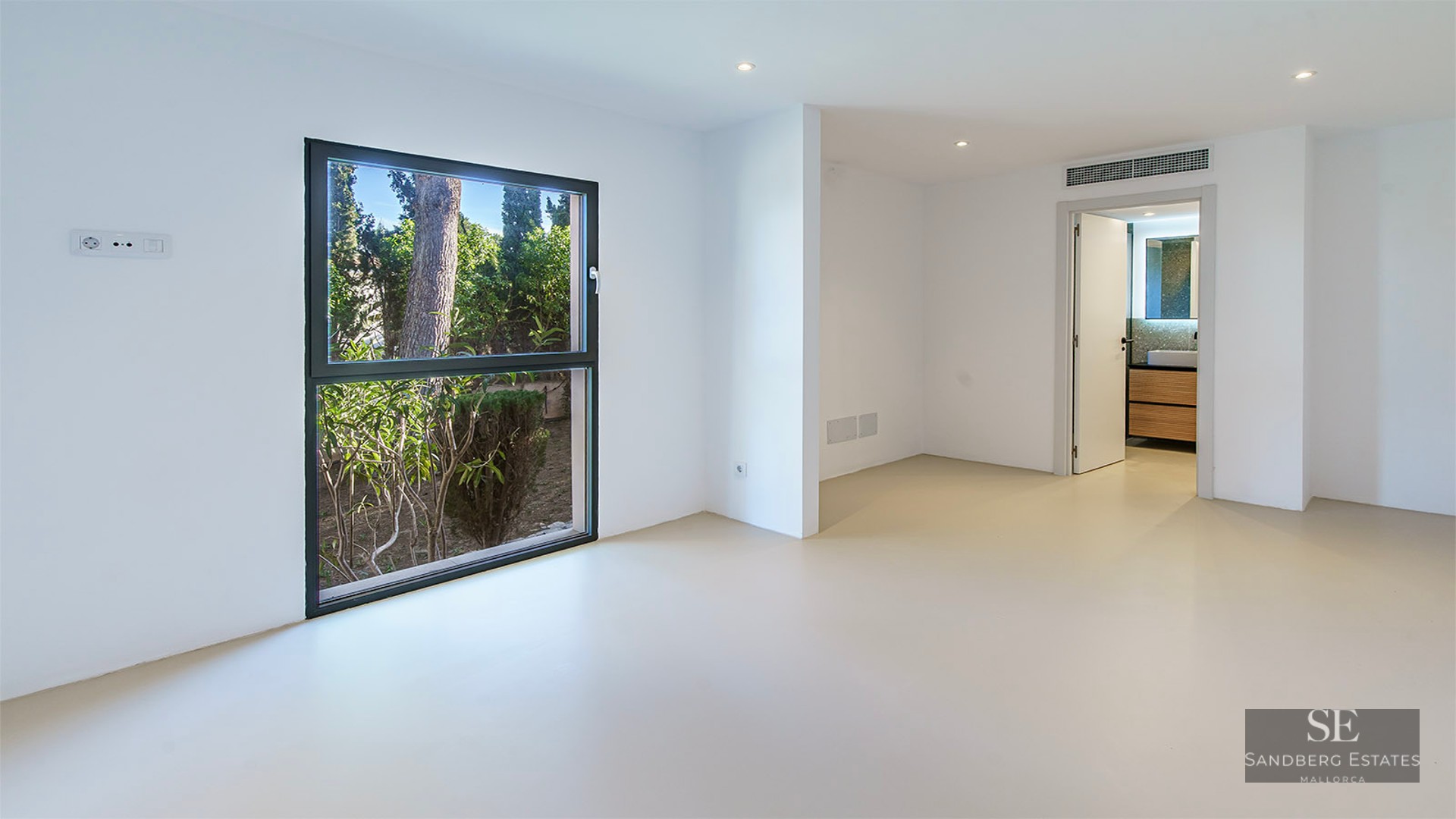 Empty minimalist bedroom with white walls, beige floors, a large garden-view window, and a door to an ensuite bathroom.