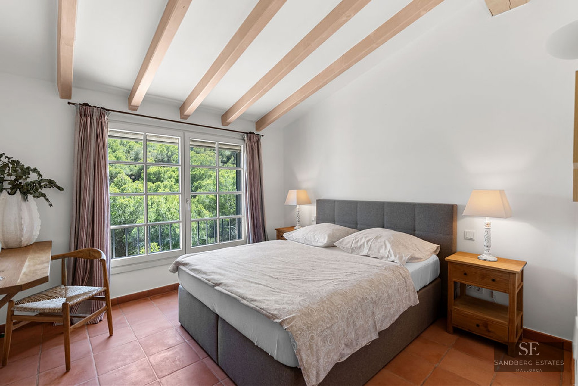 Bedroom featuring a gray double bed, exposed ceiling beams, terracotta flooring, and views of lush green trees.