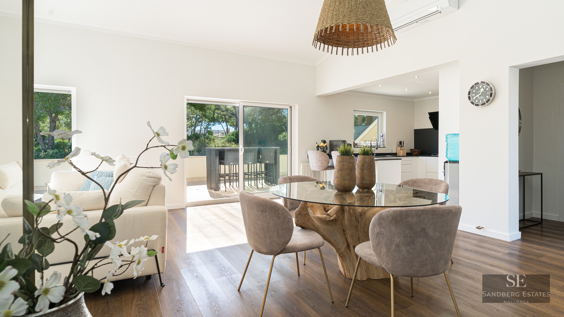 Pièce lumineuse à aire ouverte avec table à manger en verre sur socle en racine, chaises taupe et terrasse ensoleillée.