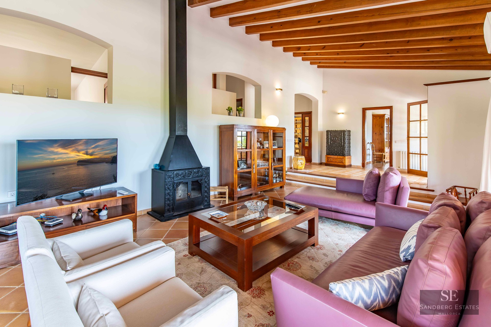 Living room featuring purple and white sofas, a central coffee table, a black fireplace, and exposed wooden ceiling beams.