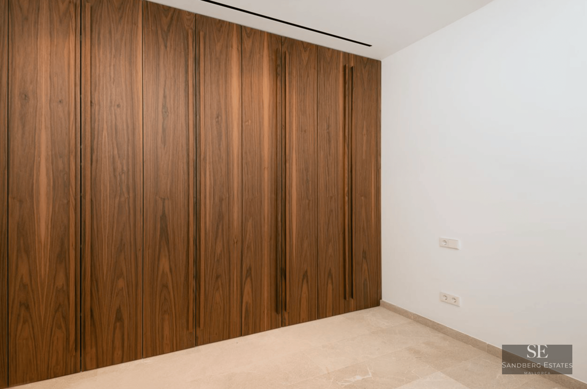 Floor-to-ceiling dark wood built-in wardrobe with vertical integrated handles in a bright room with stone floors.