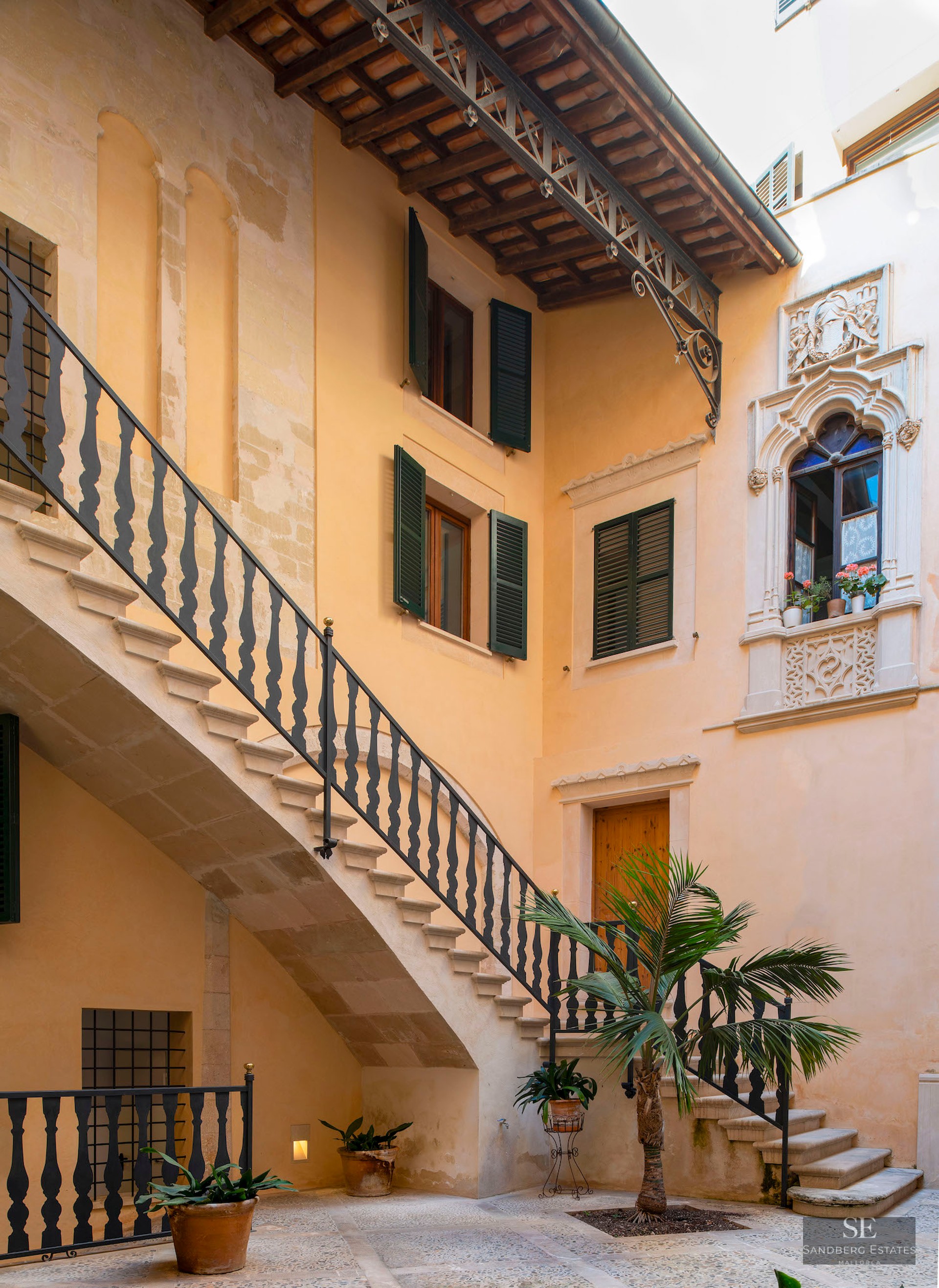 Interior courtyard featuring a stone staircase, wrought iron railings, green shutters, and a central palm tree.