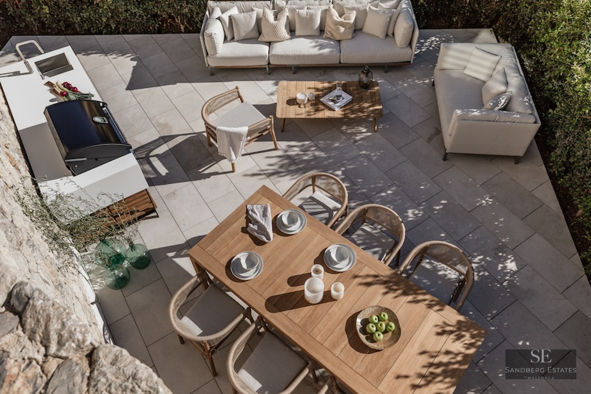 Overhead view of a modern stone patio featuring a teak dining set, white sofas, and a sleek outdoor kitchen with BBQ.