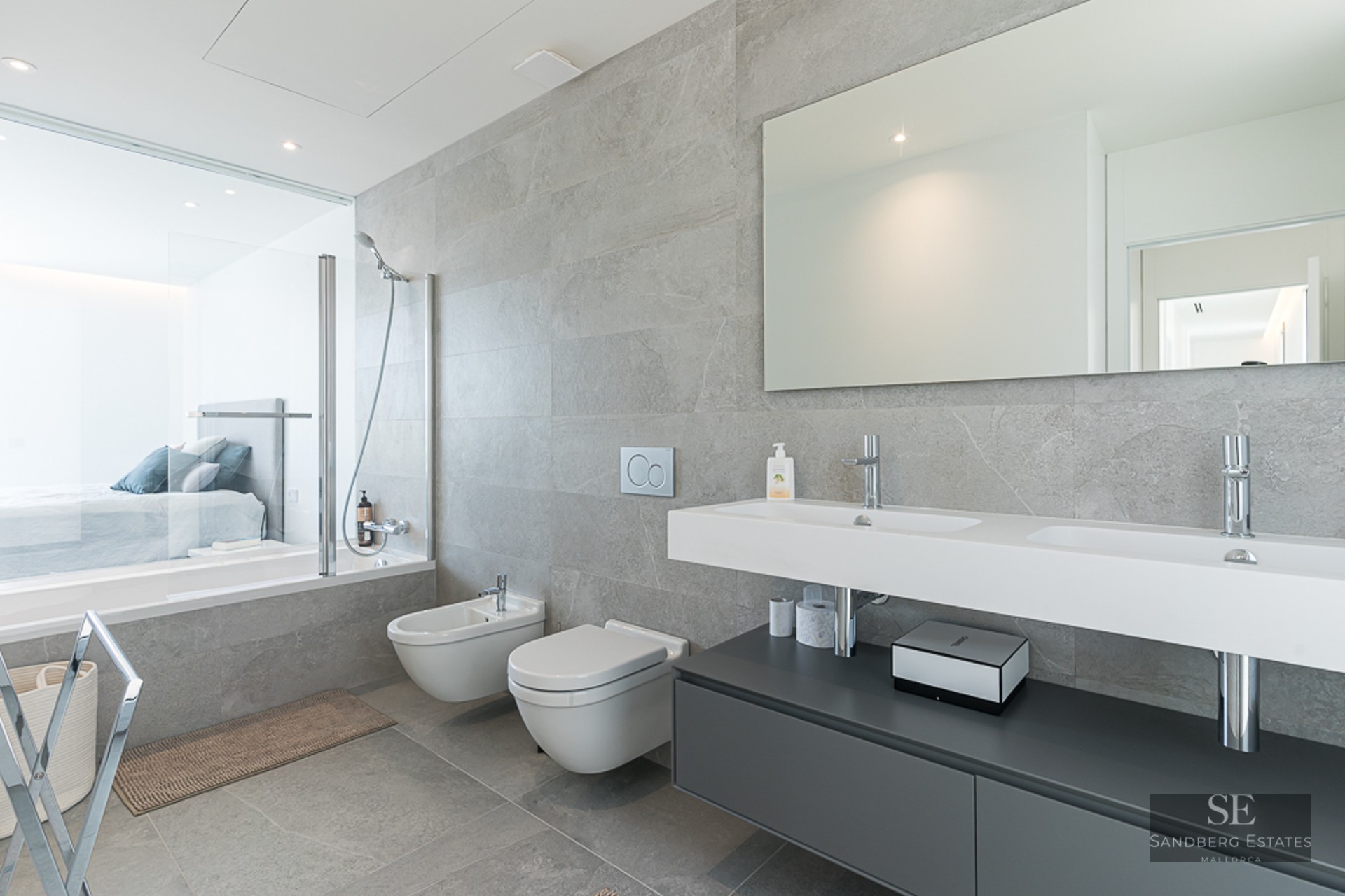 Modern bathroom featuring grey stone tiles, double vanity, wall-hung toilet, and a glass wall looking into a bedroom.