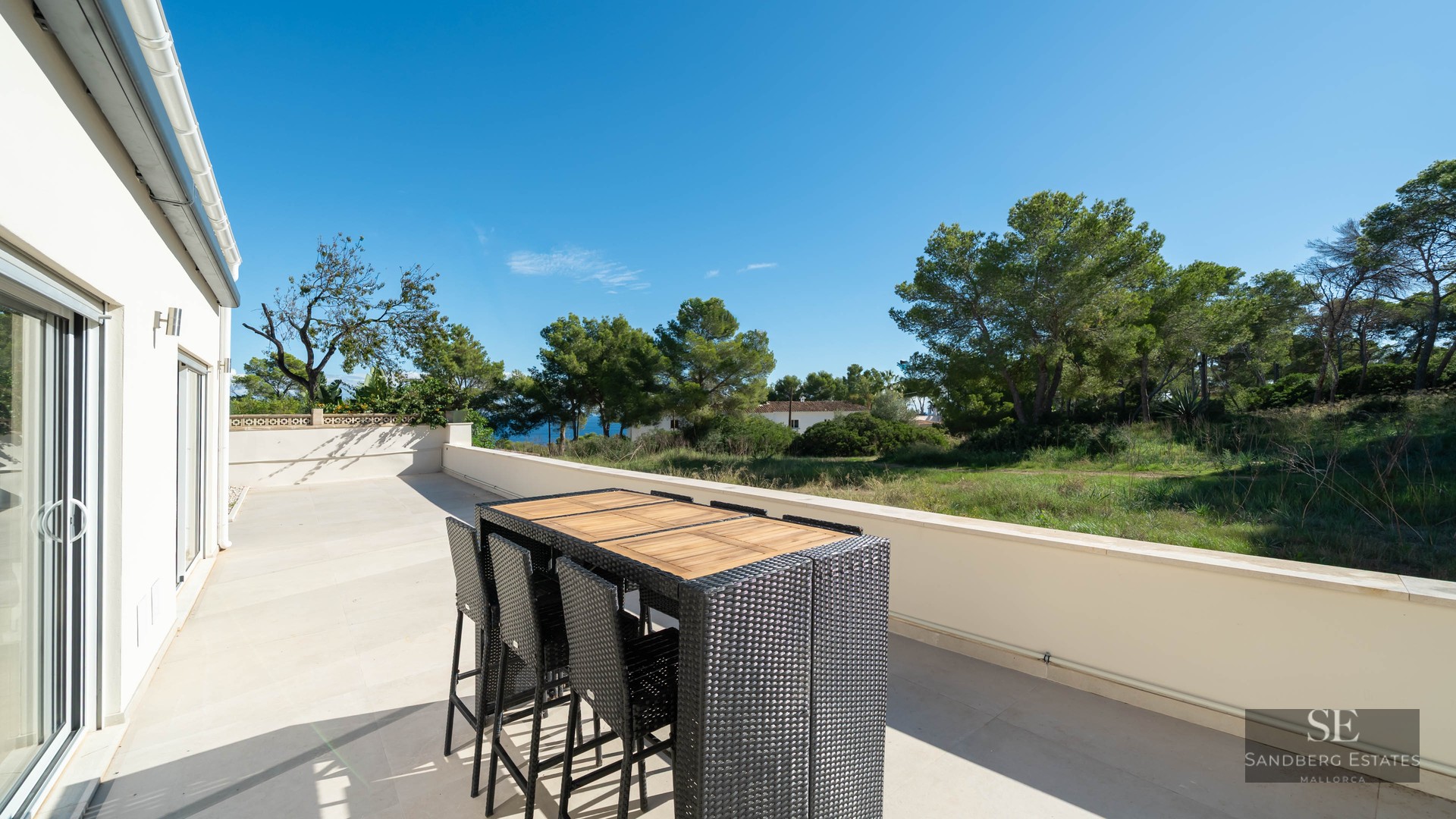 Terrasse ensoleillée avec table haute en rotin et vue sur les pins et la mer sous un ciel bleu dégagé.