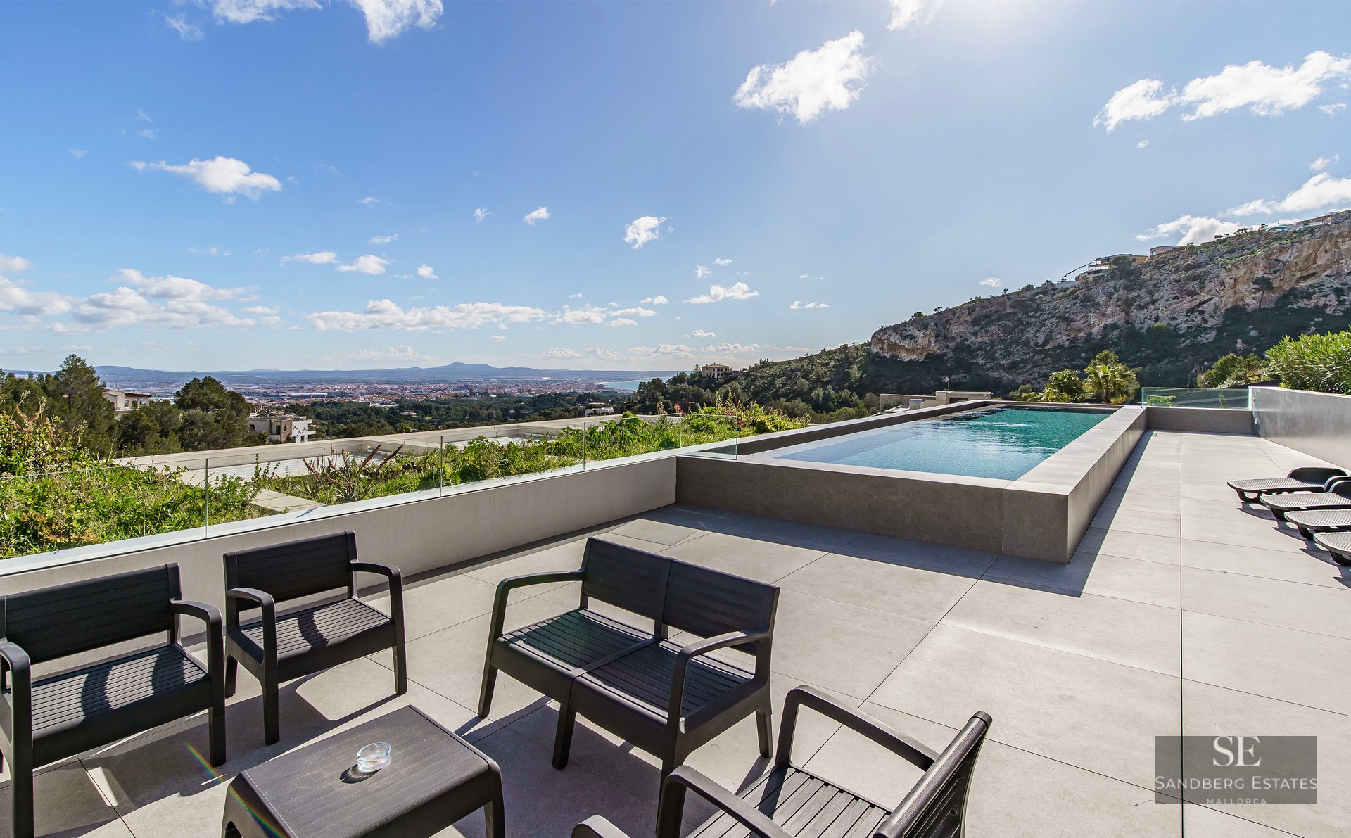 Luxury terrace featuring an infinity pool, glass railings, black outdoor furniture, and a panoramic city and sea view.