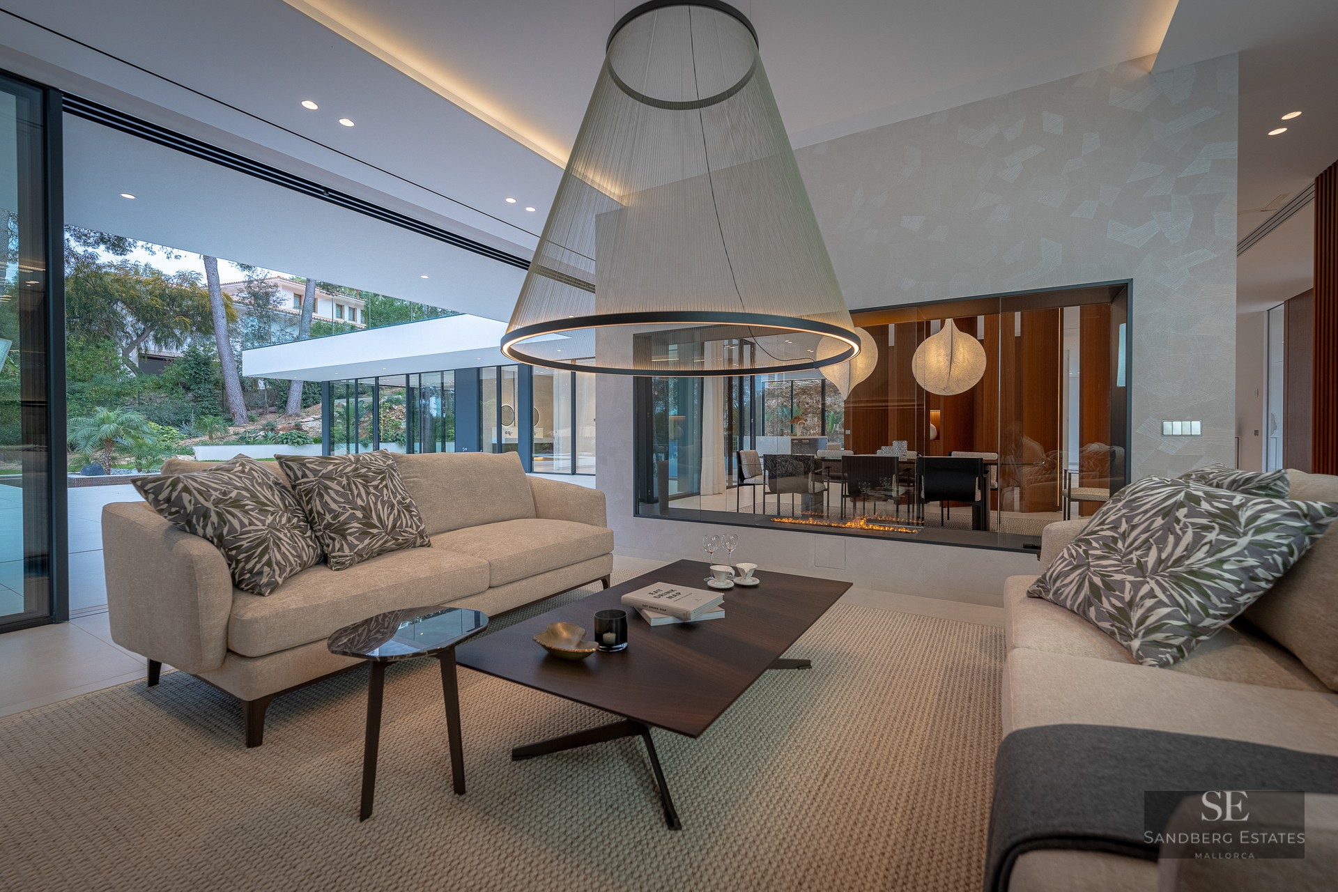 Elegant living room with beige sofas, large conical pendant light, glass fireplace, and floor-to-ceiling garden views.