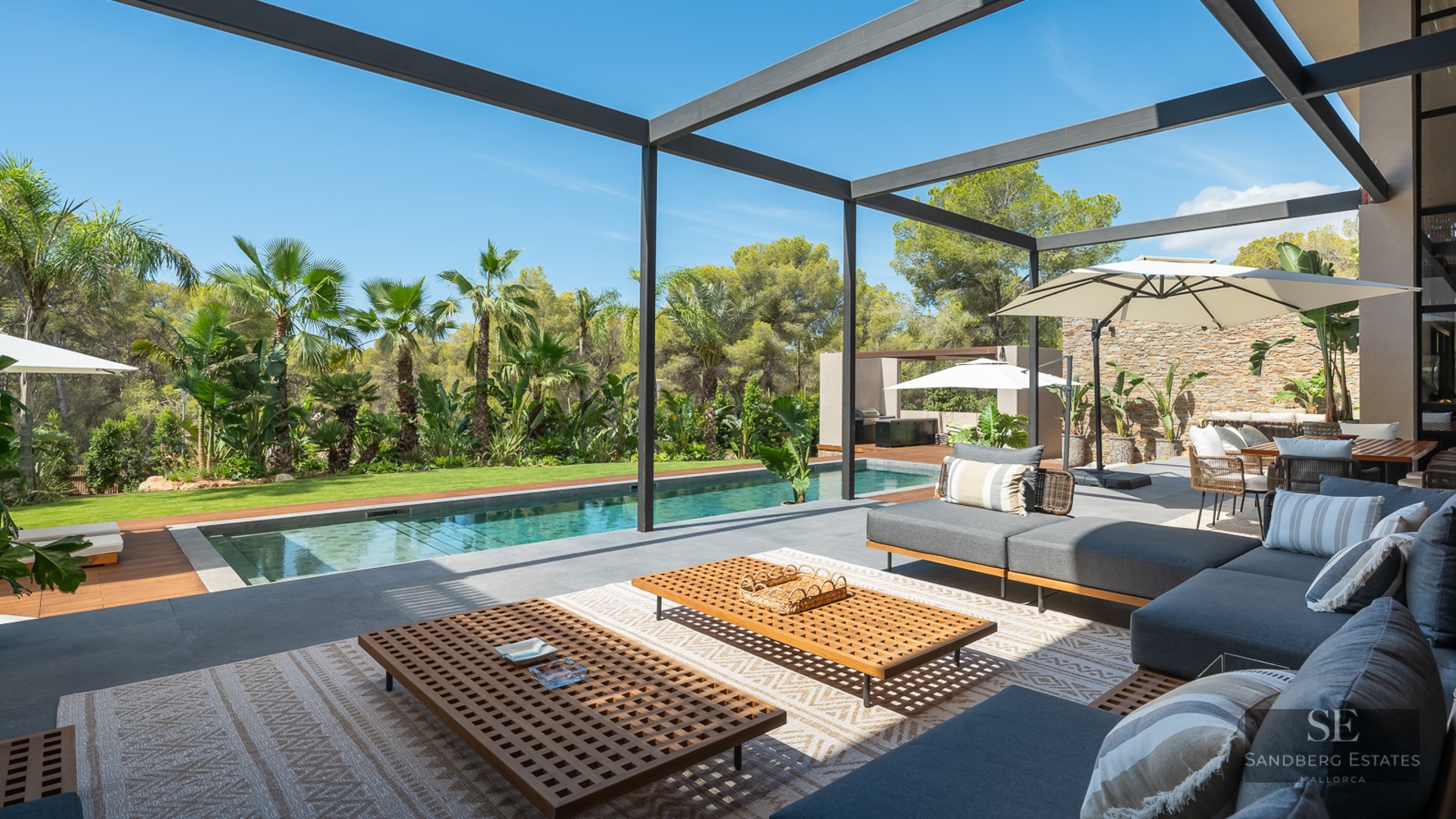 Modern outdoor lounge with grey sofas and wooden tables next to a swimming pool and lush tropical garden.