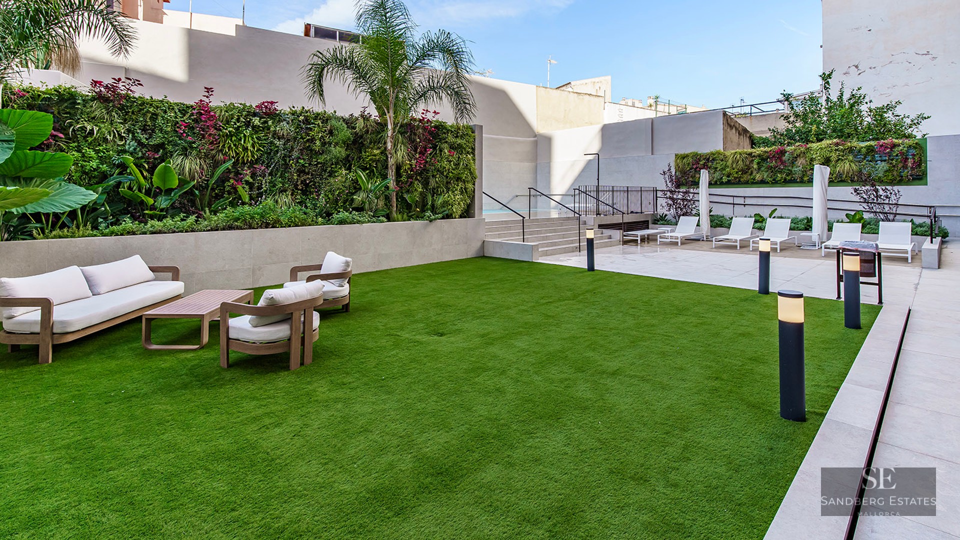 Modern outdoor lounge area with white sofas on a green lawn next to a vertical garden and a pool deck with sun loungers.