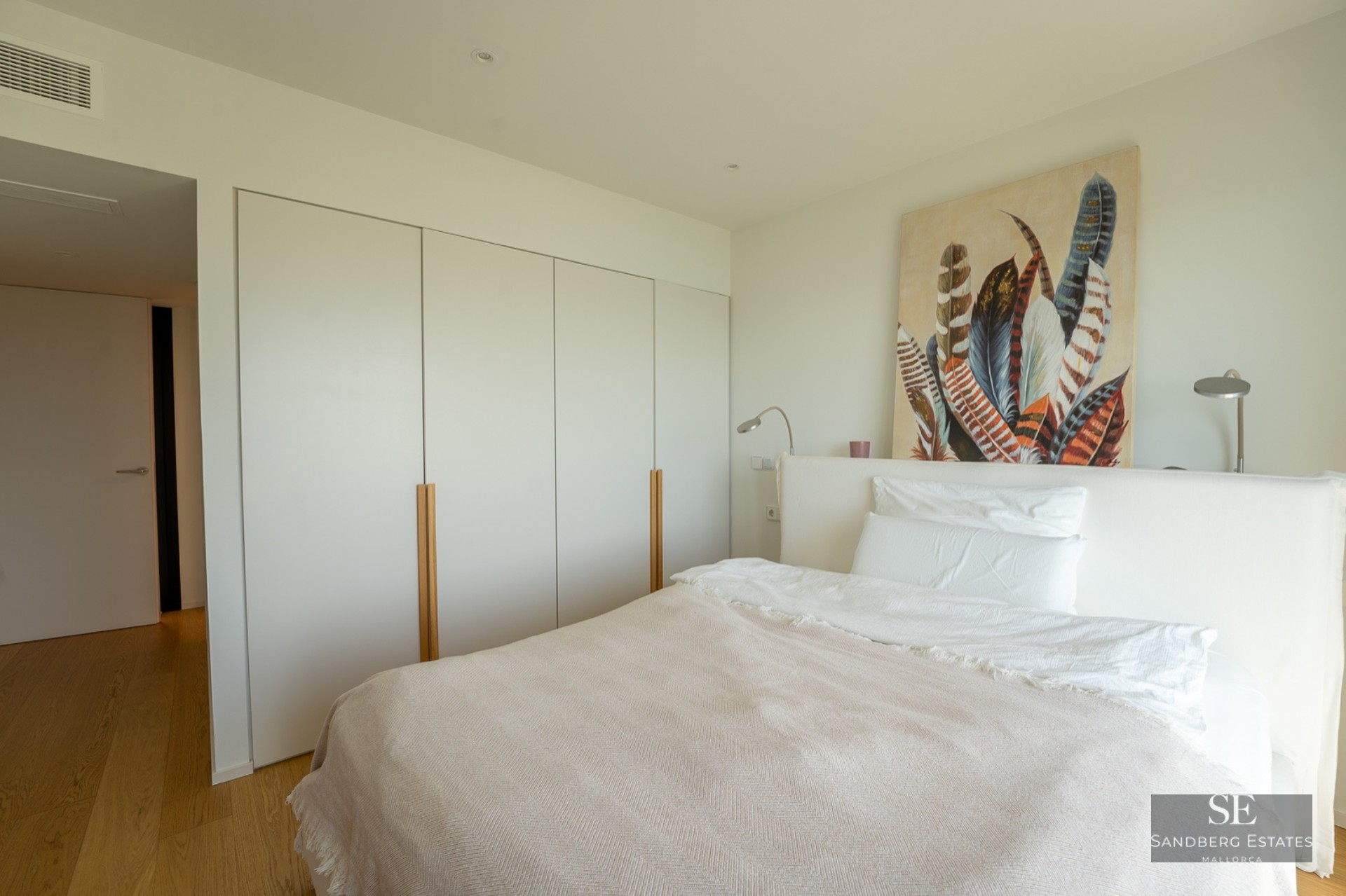 A bright bedroom featuring a white bed, large floor-to-ceiling built-in wardrobes, and feather artwork.