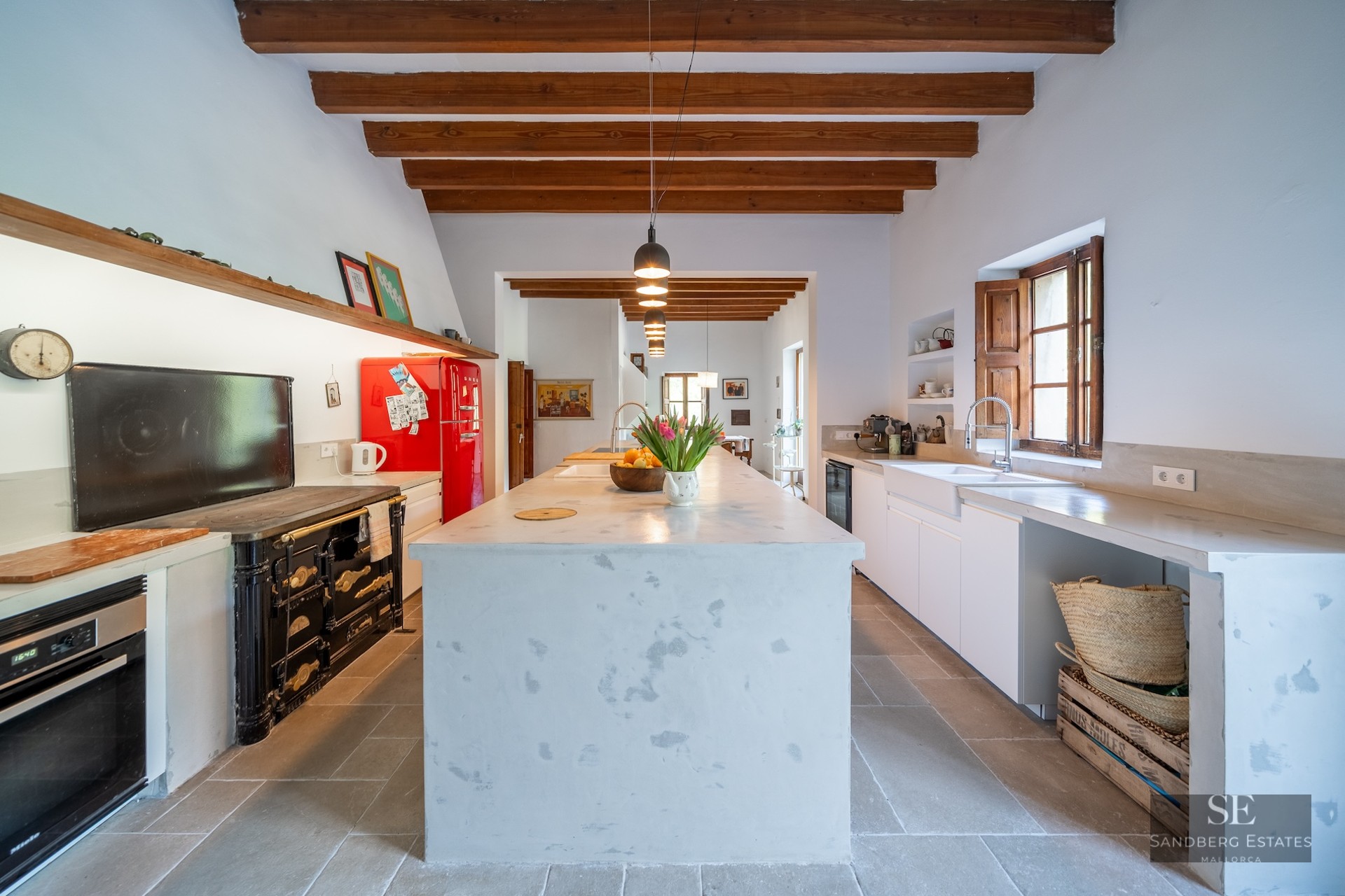 Spacious kitchen featuring a large white central island, exposed wooden beams, a retro red fridge, and a cast-iron stove.