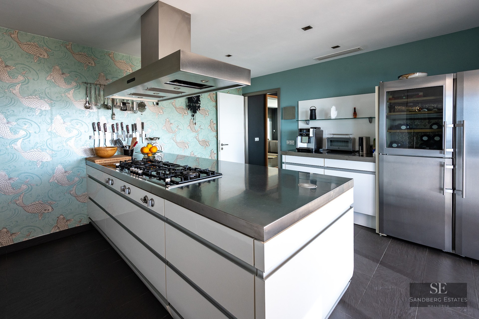A luxury kitchen featuring a central stainless steel island, fish-patterned wallpaper, and high-end appliances.
