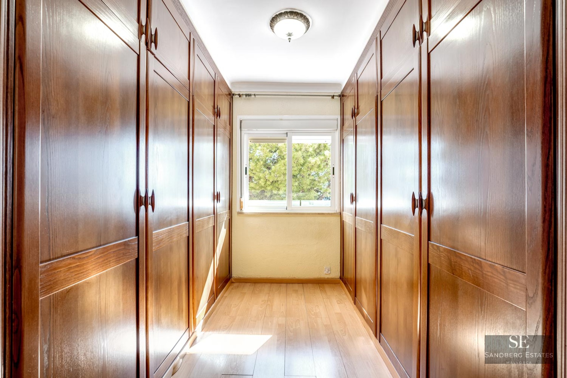 Hallway with large built-in wooden wardrobes on both sides and a window at the end looking out to trees.