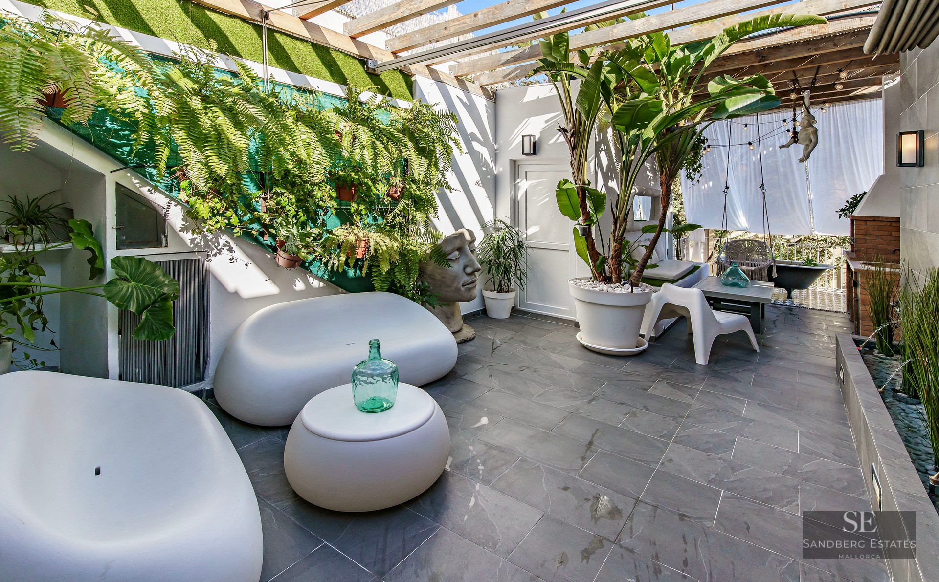Outdoor terrace featuring white curved sofas, lush green plants, gray stone flooring, and a wooden pergola.