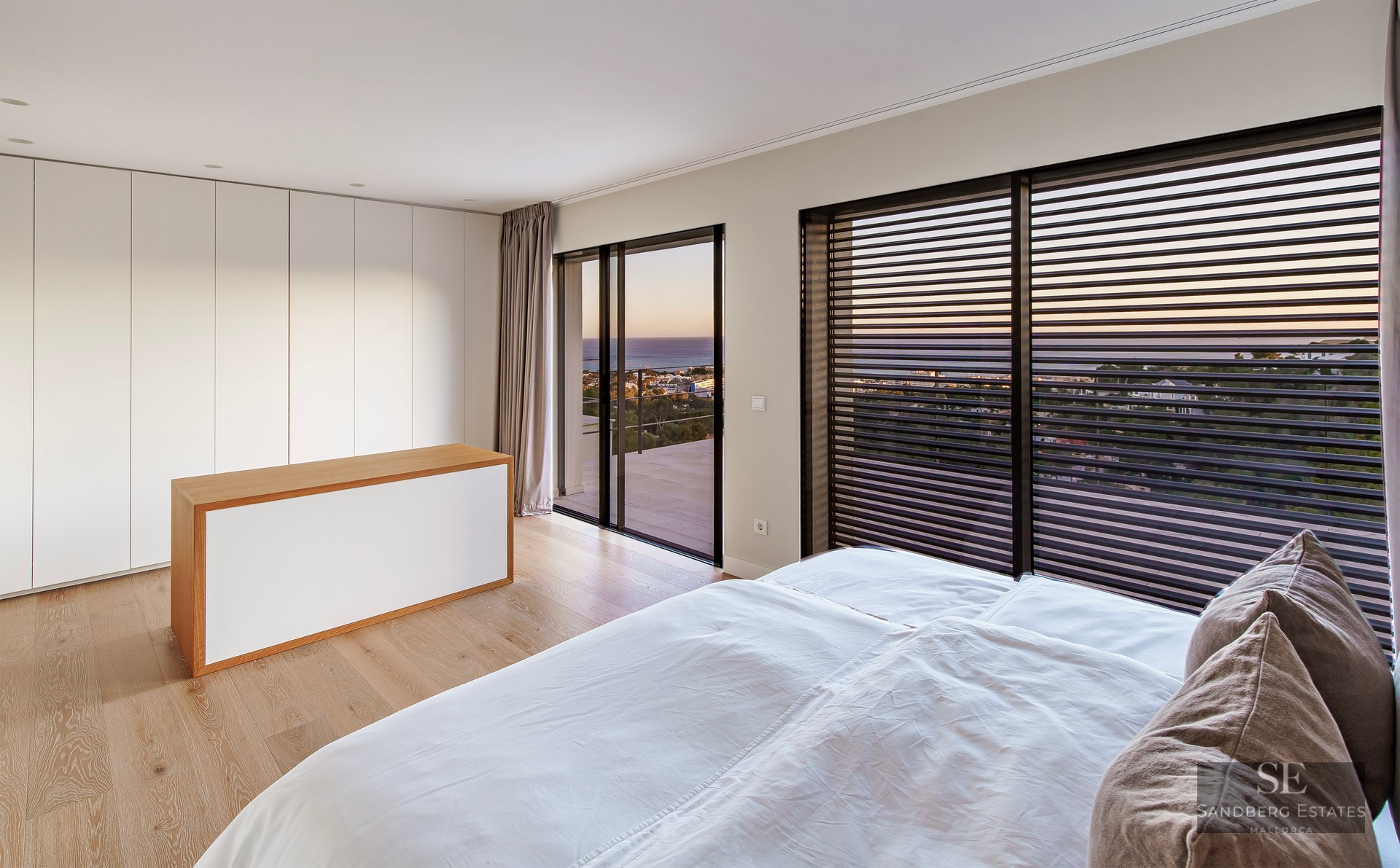 Modern bedroom with white bedding, wood floors, and large windows overlooking the sea at sunset.