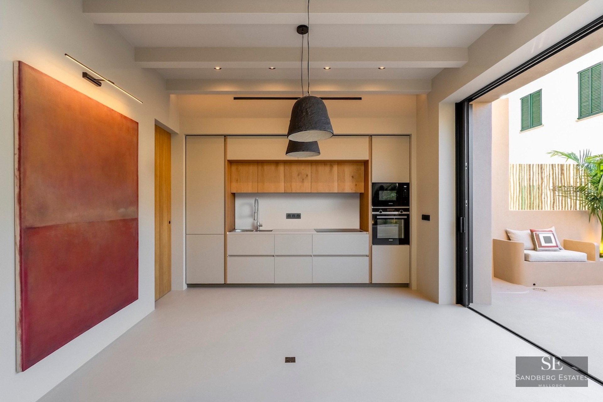 Cuisine minimaliste avec accents en bois clair, grand tableau rouge et baies vitrées ouvrant sur un patio ensoleillé.