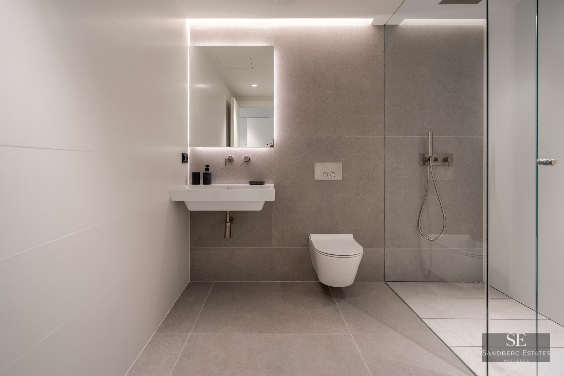 Modern bathroom featuring grey tiles, a backlit mirror above a white sink, a wall-hung toilet, and a glass shower partition.