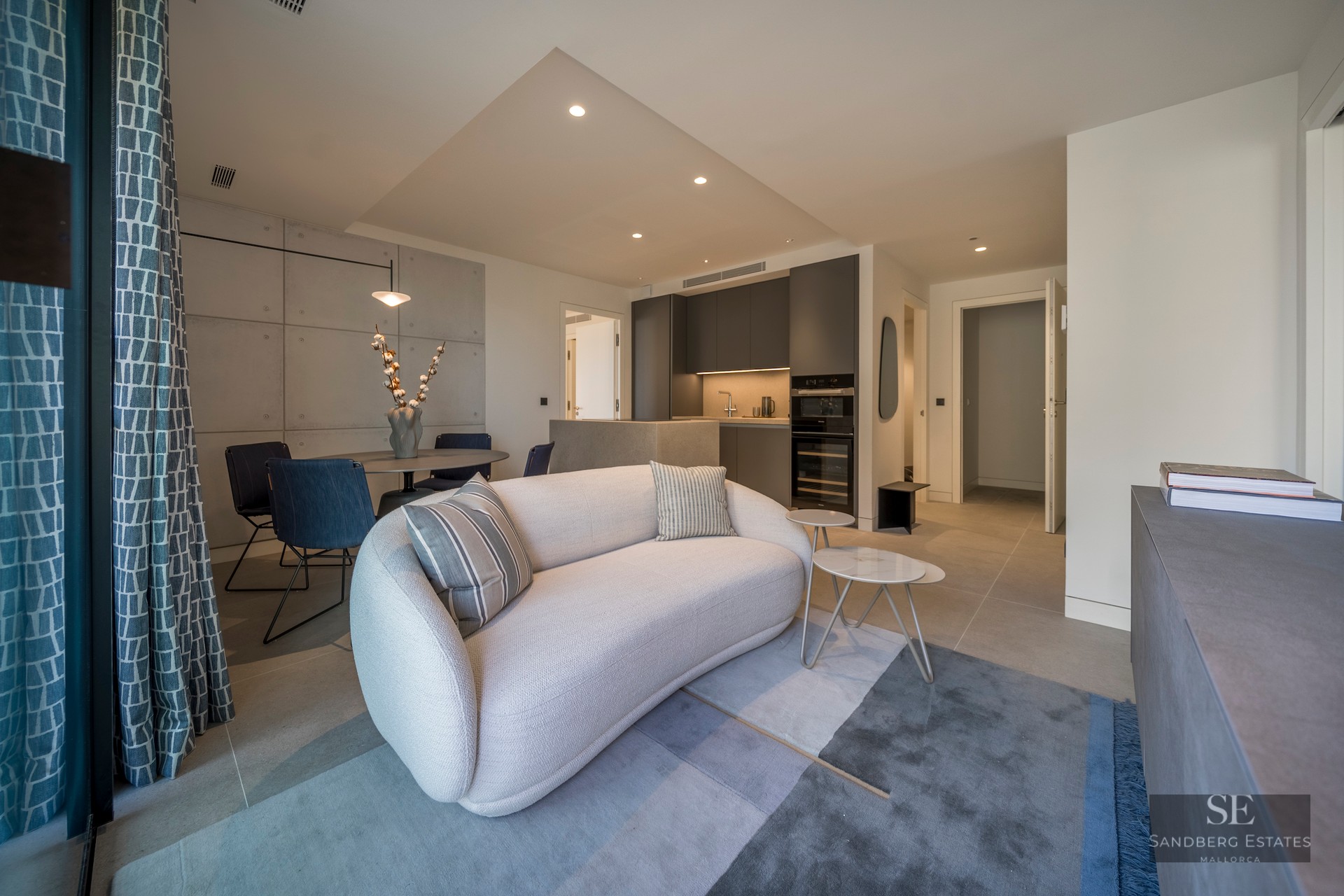 Modern living room with a curved white sofa, blue rug, and dining table, adjacent to a dark grey kitchen.