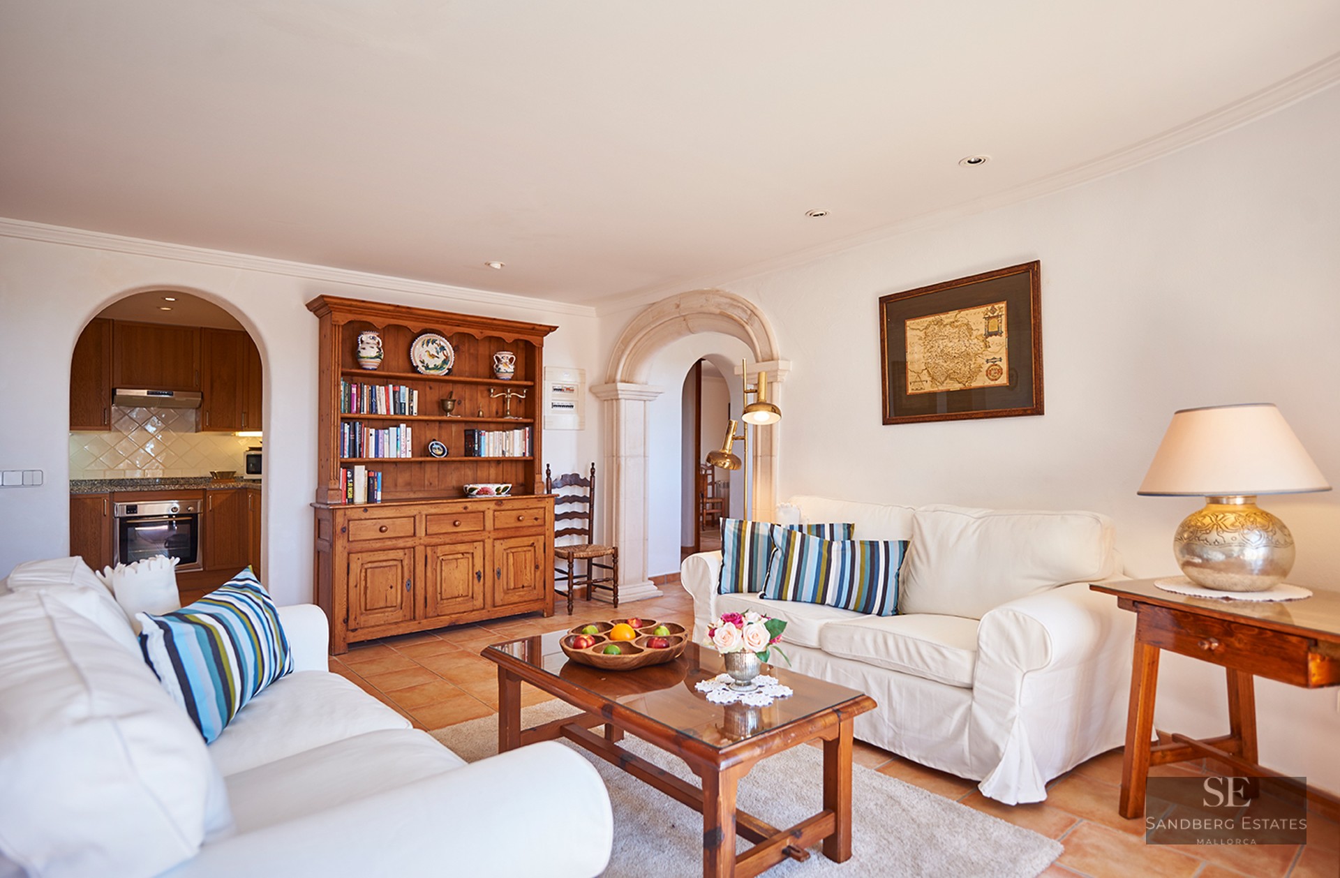 Bright living room featuring white sofas, terracotta tile floors, wooden furniture, and stone arches leading to a kitchen.