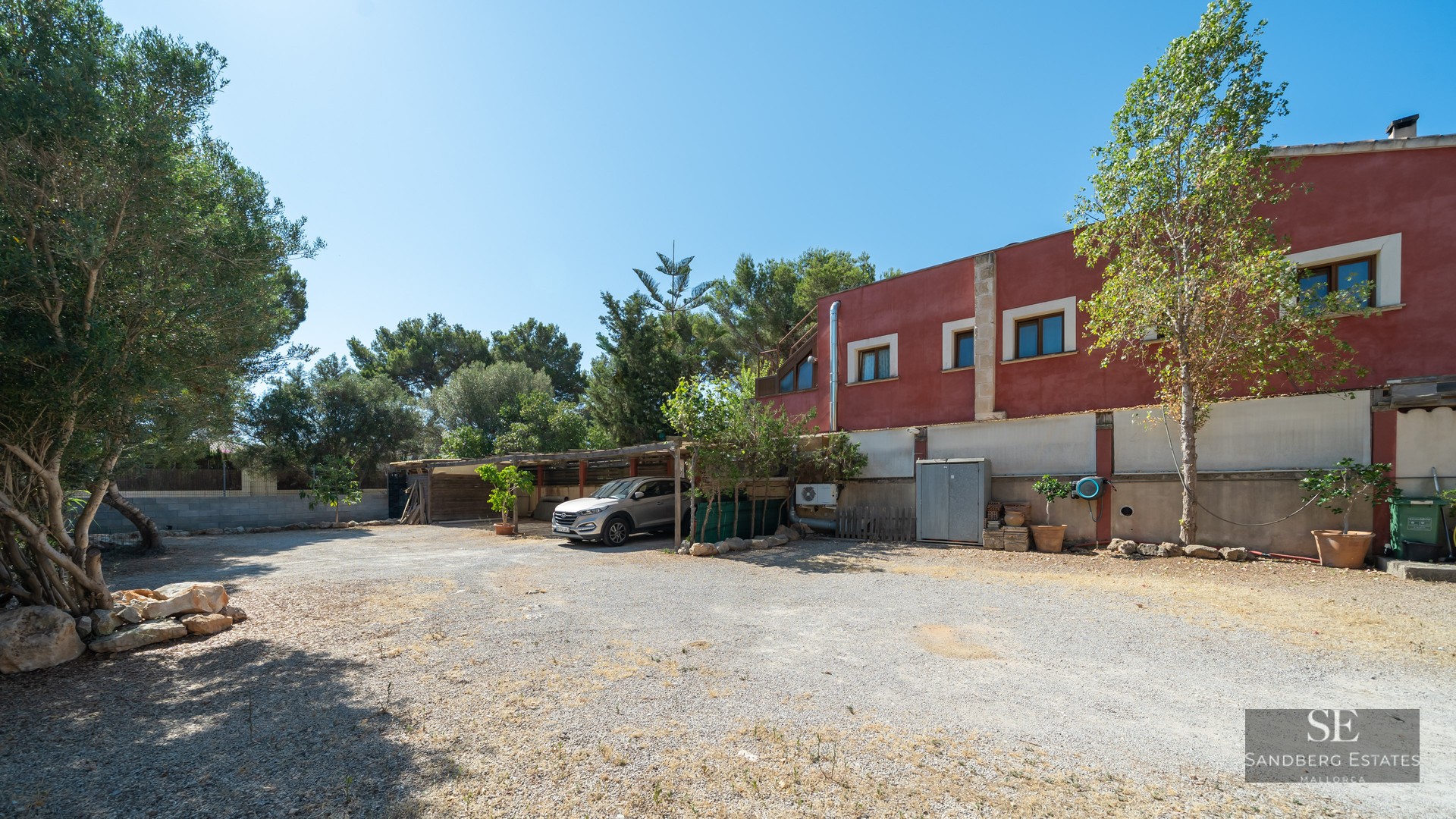 A wide gravel parking area in front of a red building with a silver car parked under a wooden carport.