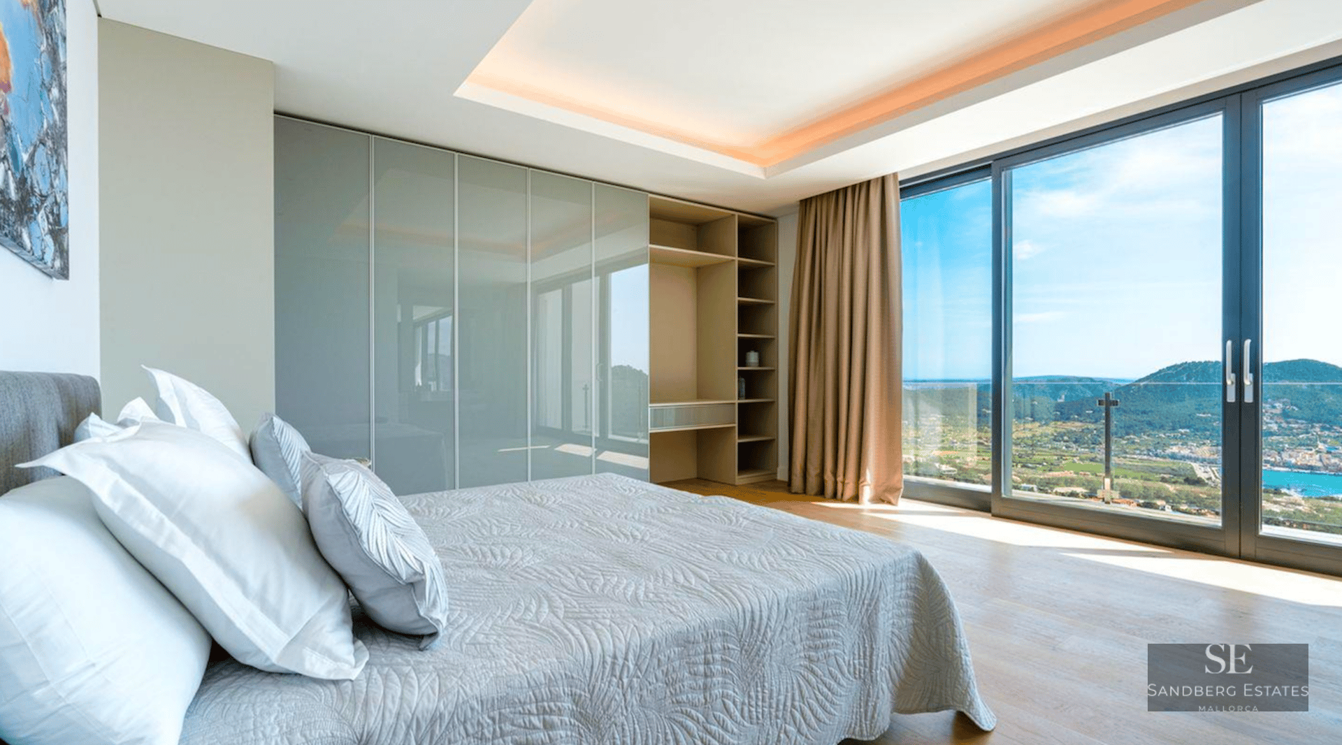 Modern bedroom with large floor-to-ceiling windows overlooking a coastal bay, featuring built-in closets and wooden floors.