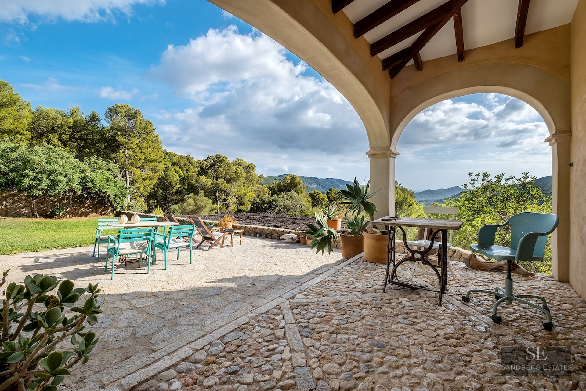 Stone terrace with arches overlooking a lush green valley and mountains, featuring teal and wood outdoor furniture.
